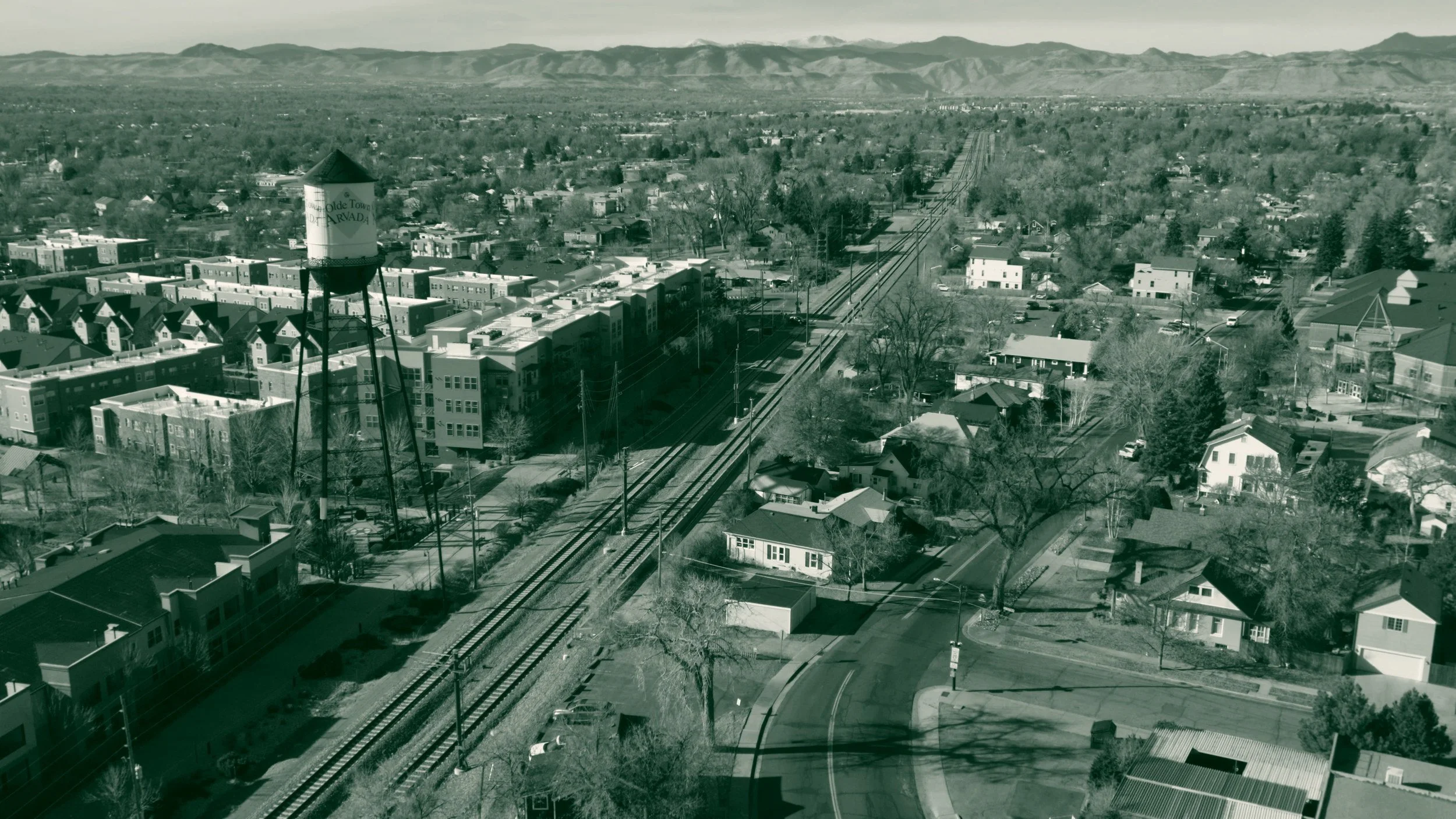 The Olde Town Arvada water tower standing above the rail corridor in Arvada, Colorado, with brick apartment buildings and single-story houses on either side and the Front Range across the horizon.