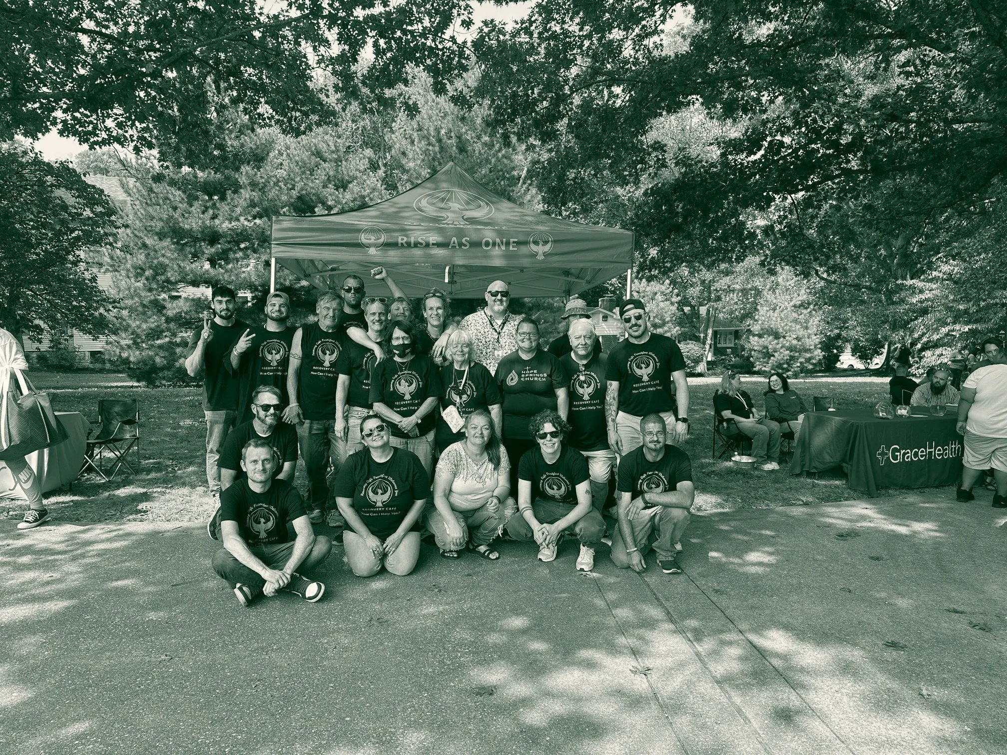 Staff and members of Recovery Café Lexington gathered for a group photograph under a Rise As One canopy at an outdoor community event.