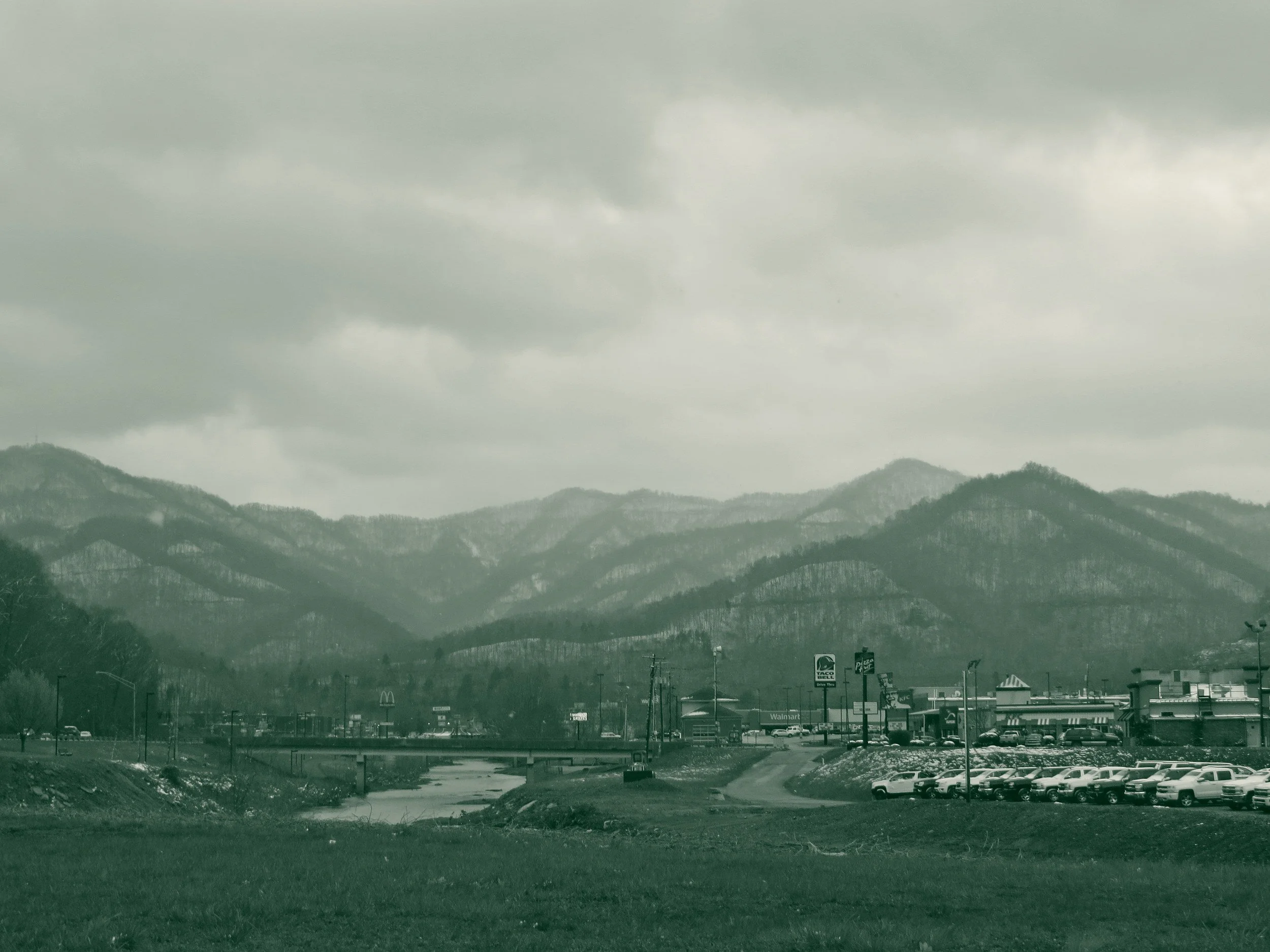 Layered Appalachian ridges under heavy cloud above a valley floor in Harlan County, Kentucky, with a creek, a bridge and a line of highway storefronts below.
