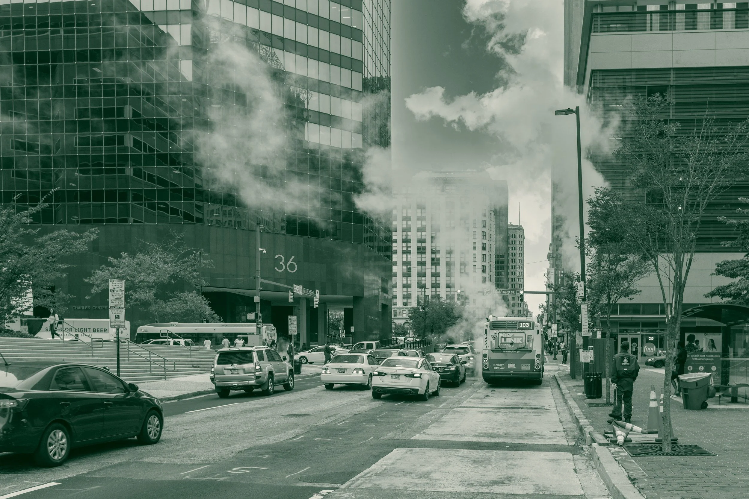 Steam rising over traffic and a city bus on Charles Street near Lombard Street in downtown Baltimore, with office towers on either side.