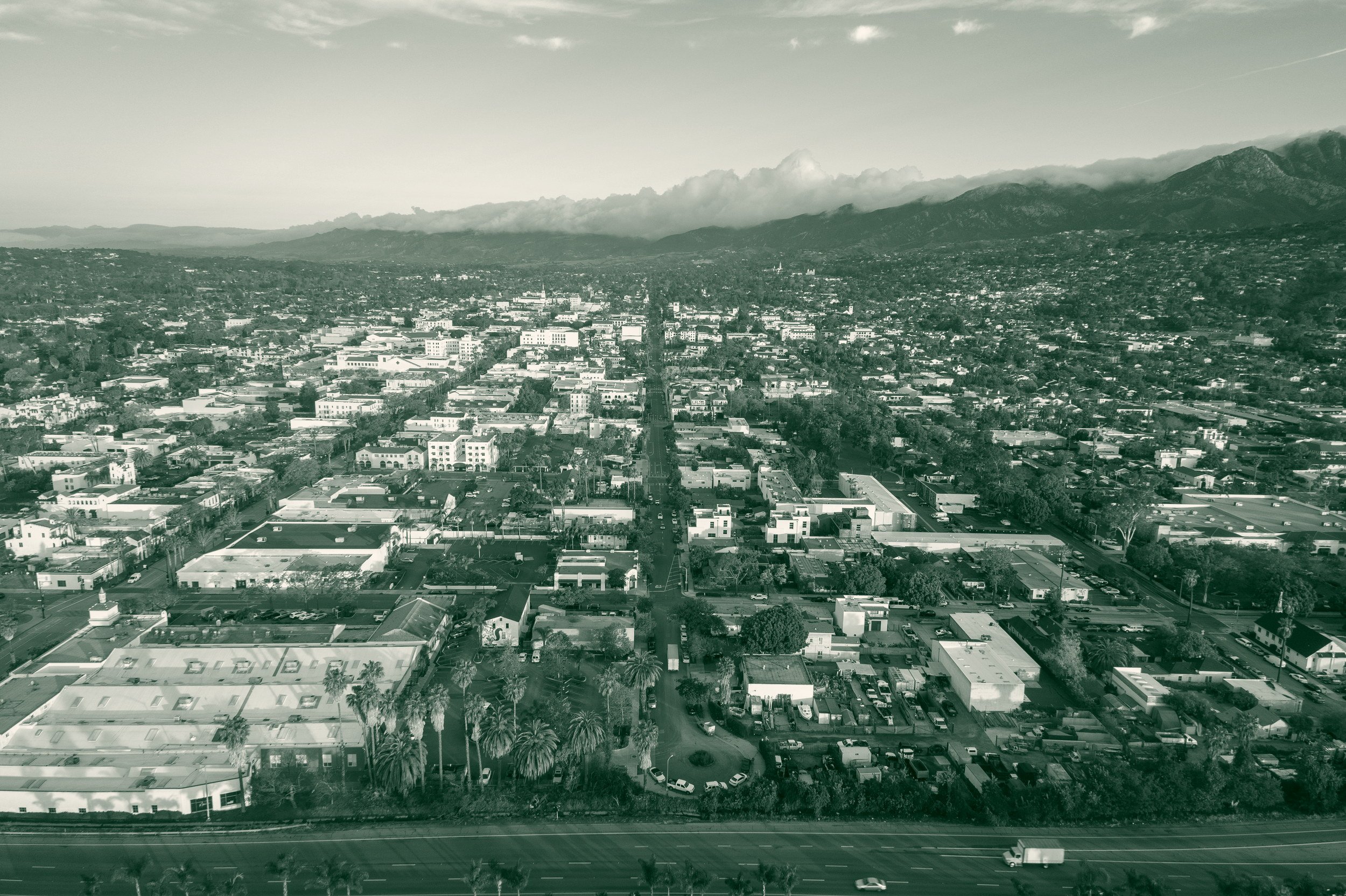 Aerial view of downtown Santa Barbara, California, looking inland along a straight street grid toward the Santa Ynez Mountains, with low clouds resting on the peaks and a freeway across the foreground.