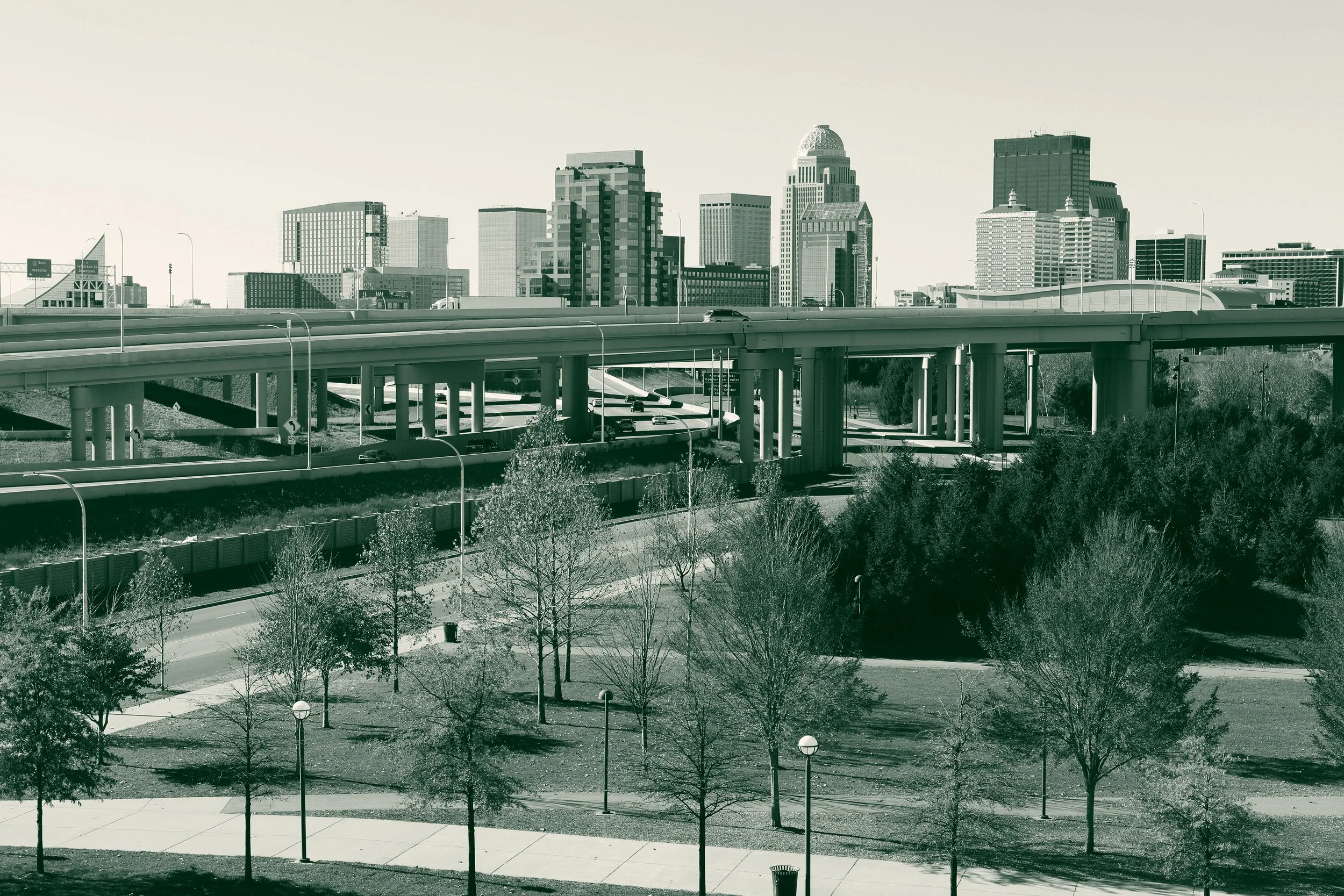 Downtown Louisville skyline behind an elevated interstate interchange, with a park, walking paths and trees in the foreground.