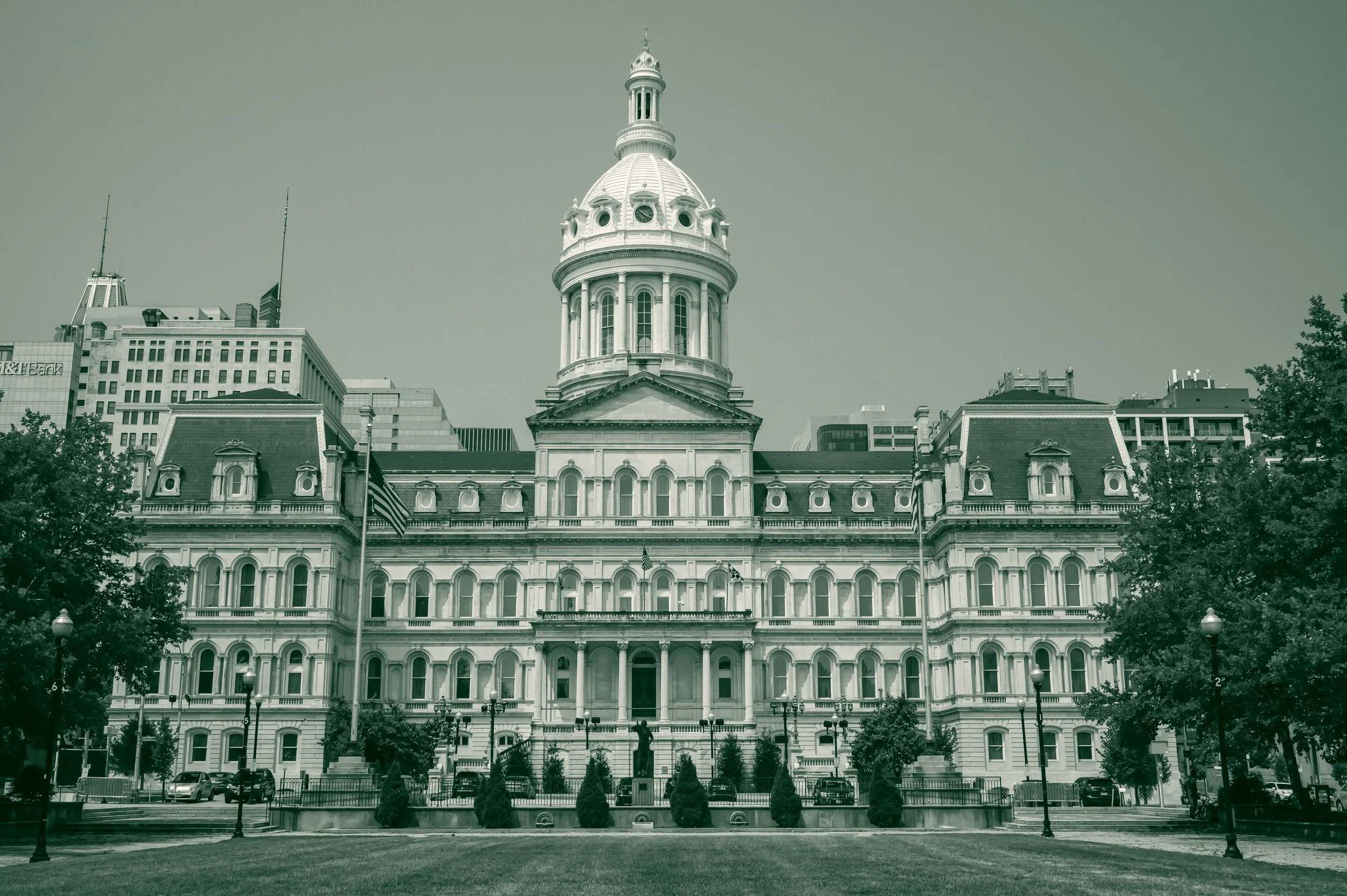 Baltimore City Hall, a white stone building with a domed tower, seen across the lawn of War Memorial Plaza with downtown office towers behind.