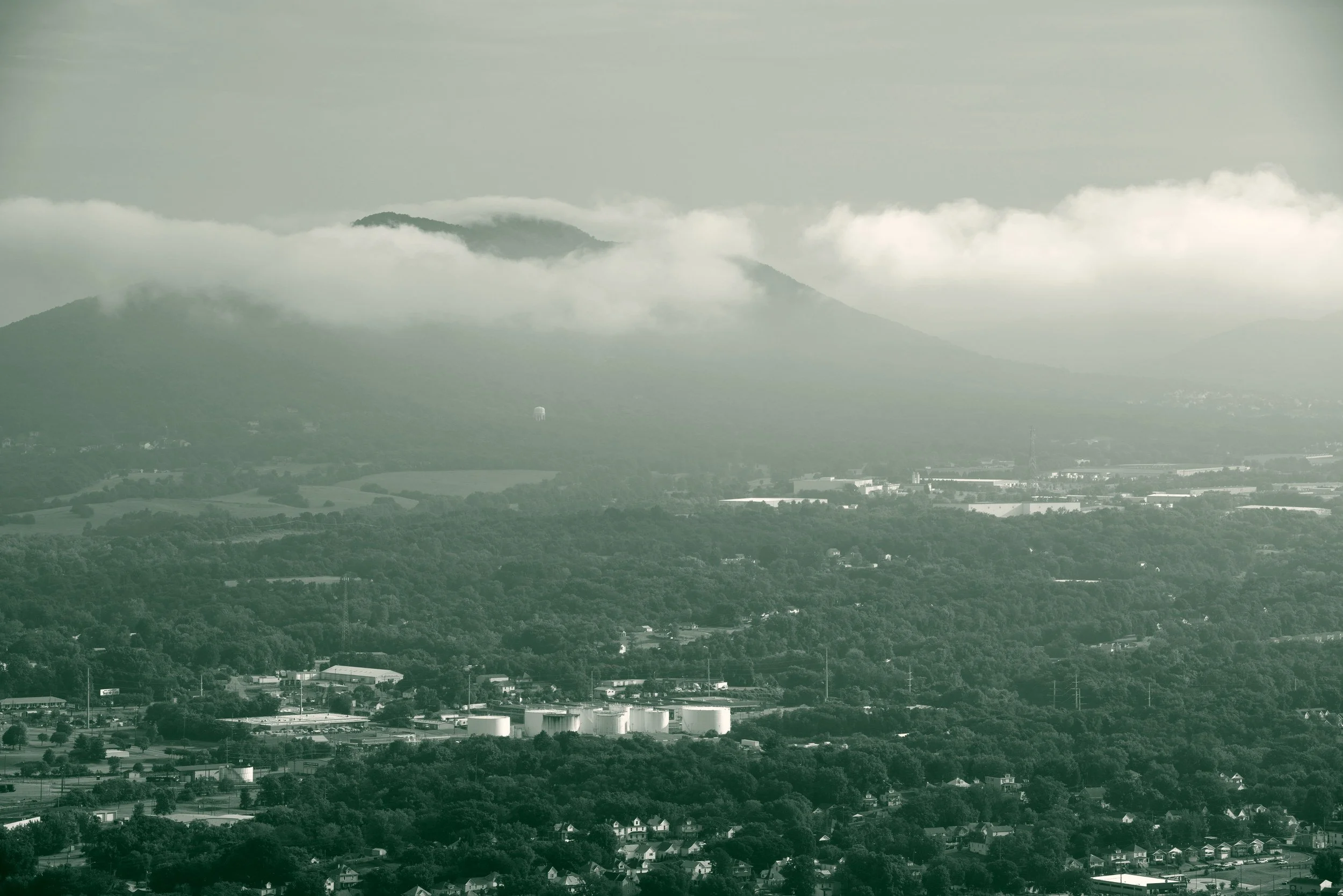 Wooded neighborhoods of Roanoke, Virginia, seen from Mill Mountain Park, with a mountain wrapped in low clouds beyond the valley.