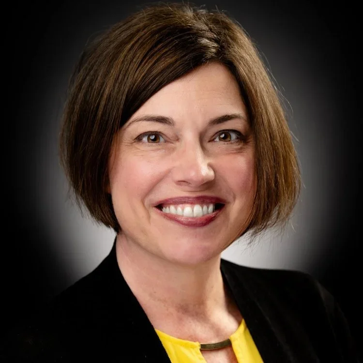 Portrait of a woman with short brown hair wearing a black blazer and yellow top, smiling against a dark background.