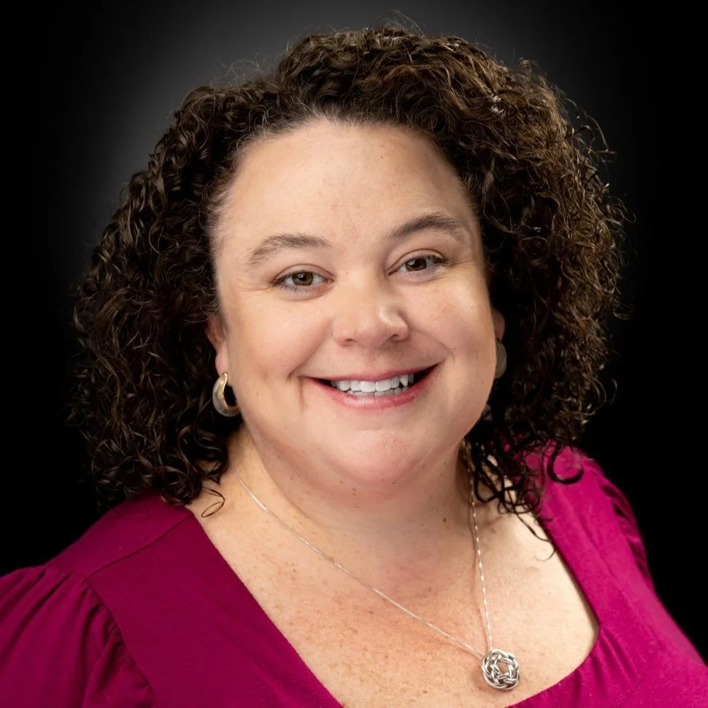 Headshot of a smiling woman with curly brown hair wearing a magenta top, hoop earrings, and a silver necklace against a black background.