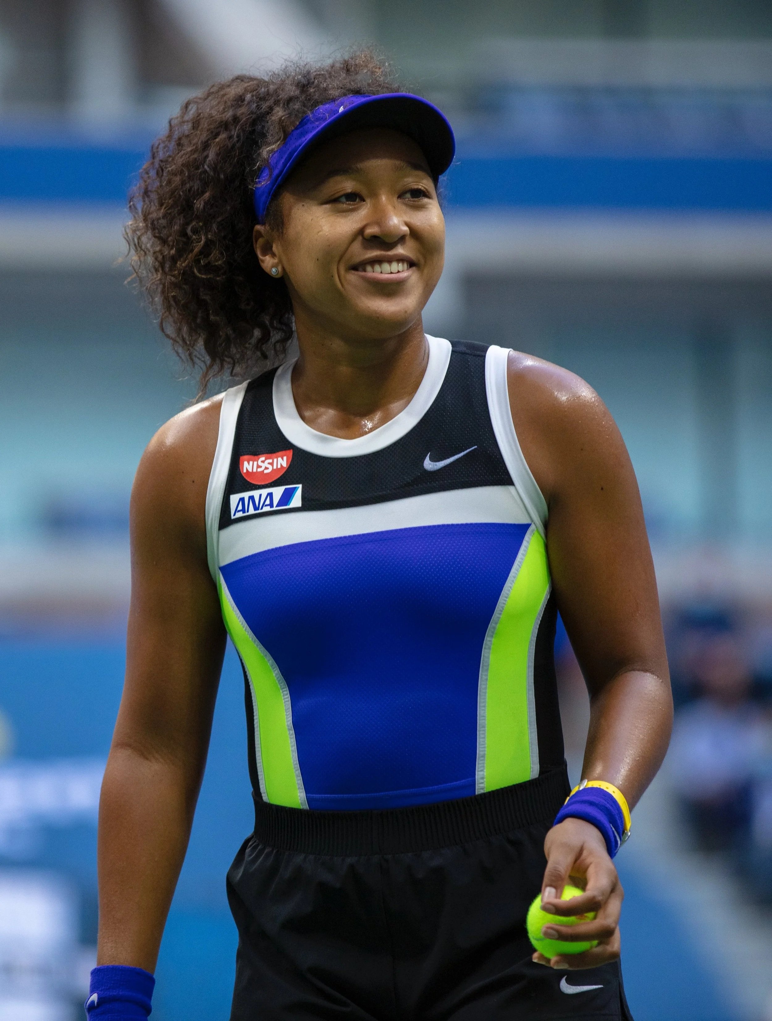 Naomi Osaka standing on a court, holding a tennis ball in her right hand, wearing her iconic sports dress with sponsor logos, a blue visor, and sports accessories. This is the match in 2018 where she be came champion of the U.S. Open Tennis Champions