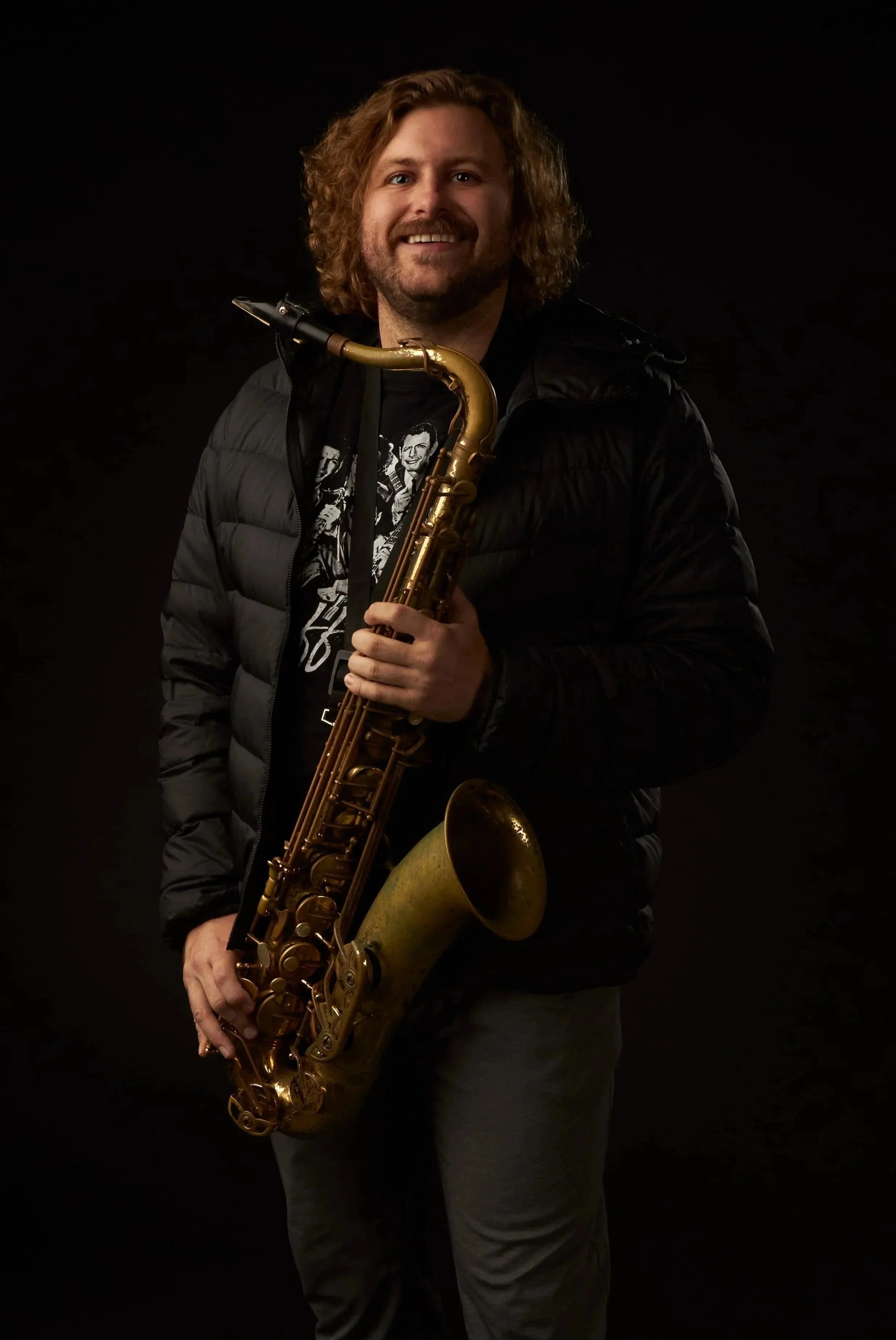 A man with long, curly hair and a beard smiling while holding a saxophone against a black background.