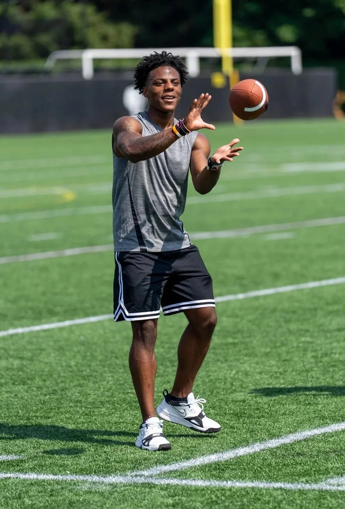 A young man wearing black shorts, a gray sleeveless sports shirt, and white Nike sneakers playing football on a grassy field.