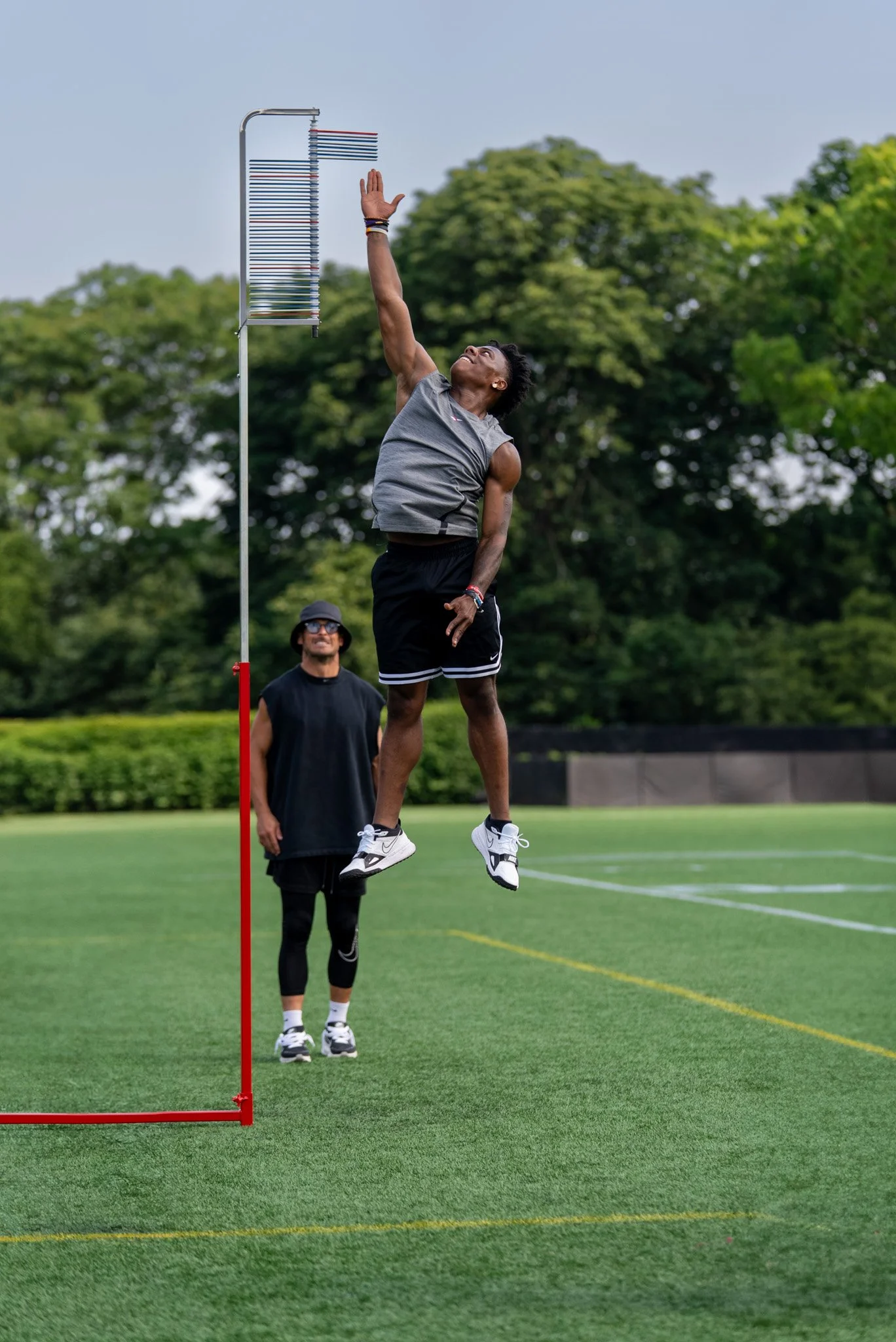 A man jumping and reaching to catch a high jump bar during an outdoor track and field event, with another man standing behind him on a green field surrounded by trees.
