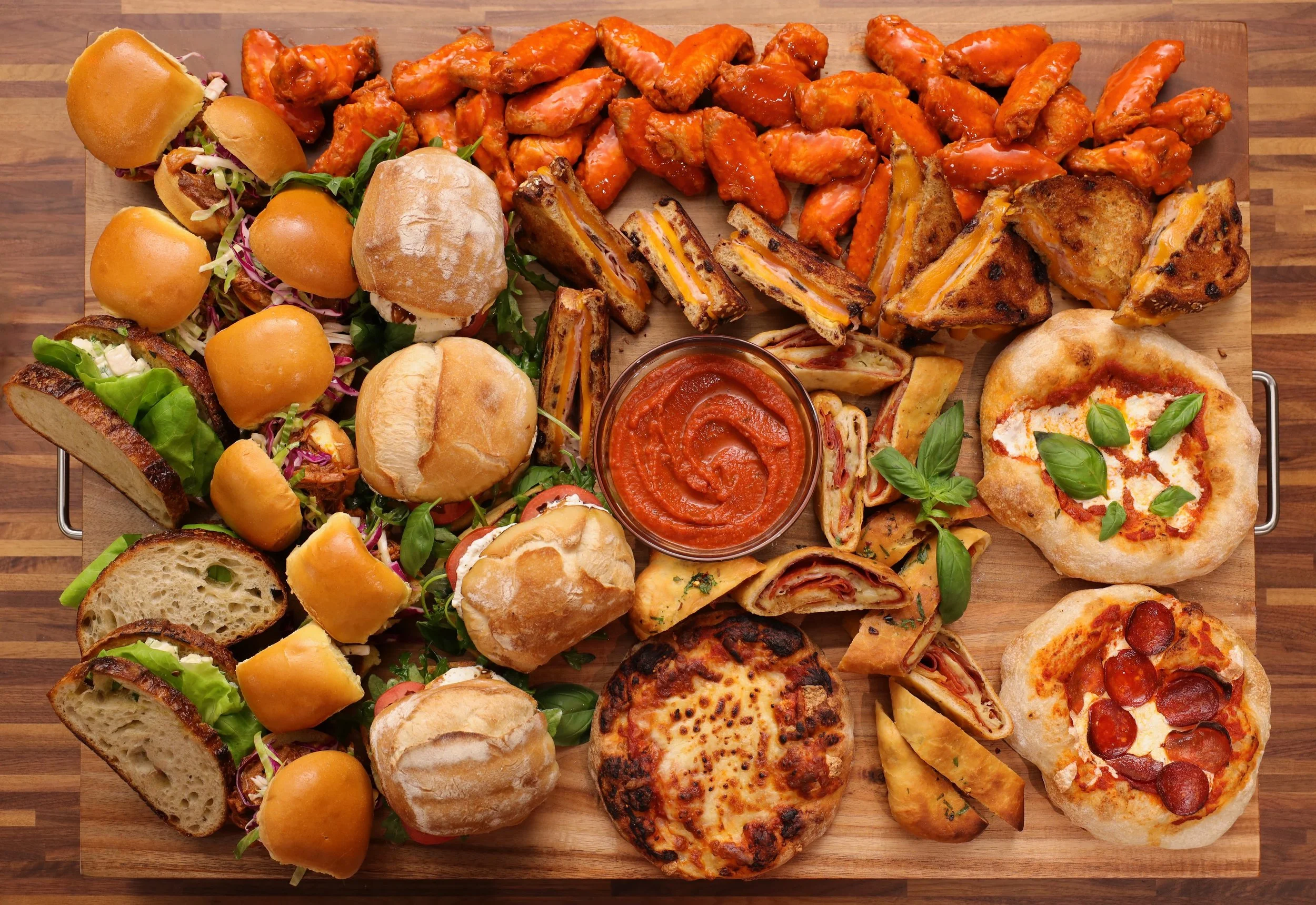 Assorted pizza slices, bread rolls, sliders, chicken wings, and a bowl of marinara sauce on a wooden serving board.
