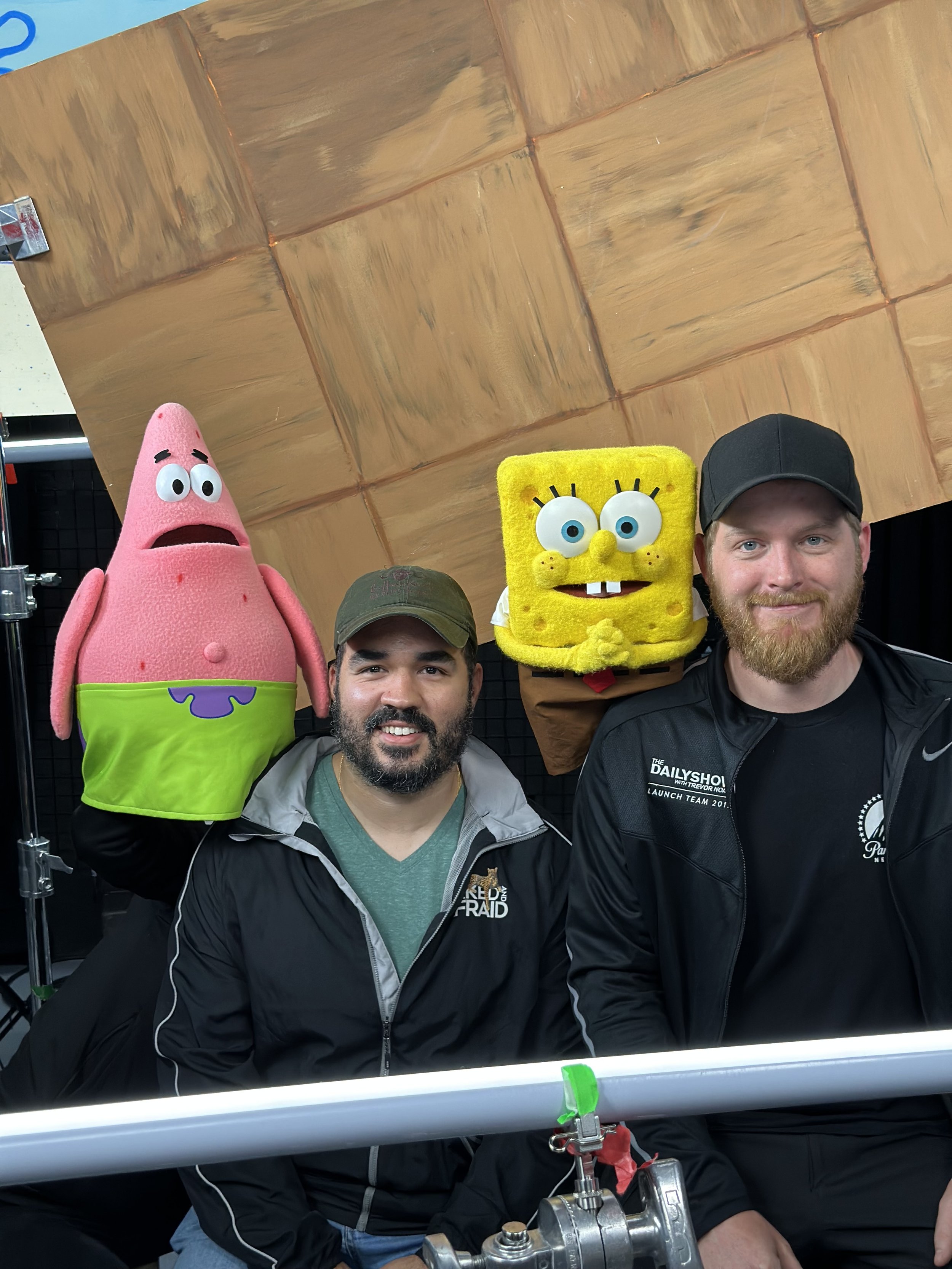 Andrew Henkelman, Derick Xavier, Patrick Star  and SpongeBob SquarePants, standing in front of a wooden wall and filming equipment during Spongebob Squarepants: Pineapple Playhouse.
