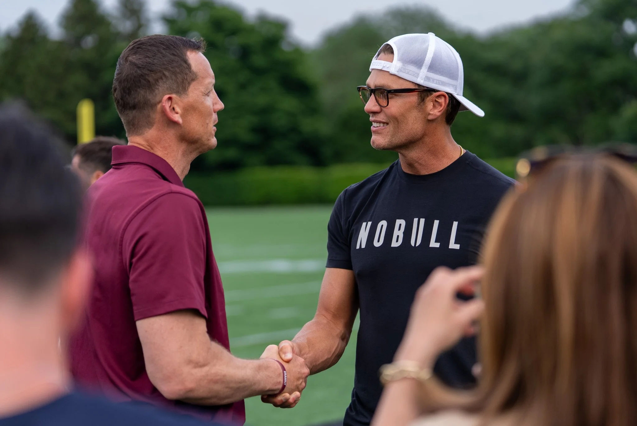 Two men shaking hands outdoors during daytime, one wearing a maroon shirt and the other wearing a black shirt with glasses and a white cap worn backwards, with green trees in the background.