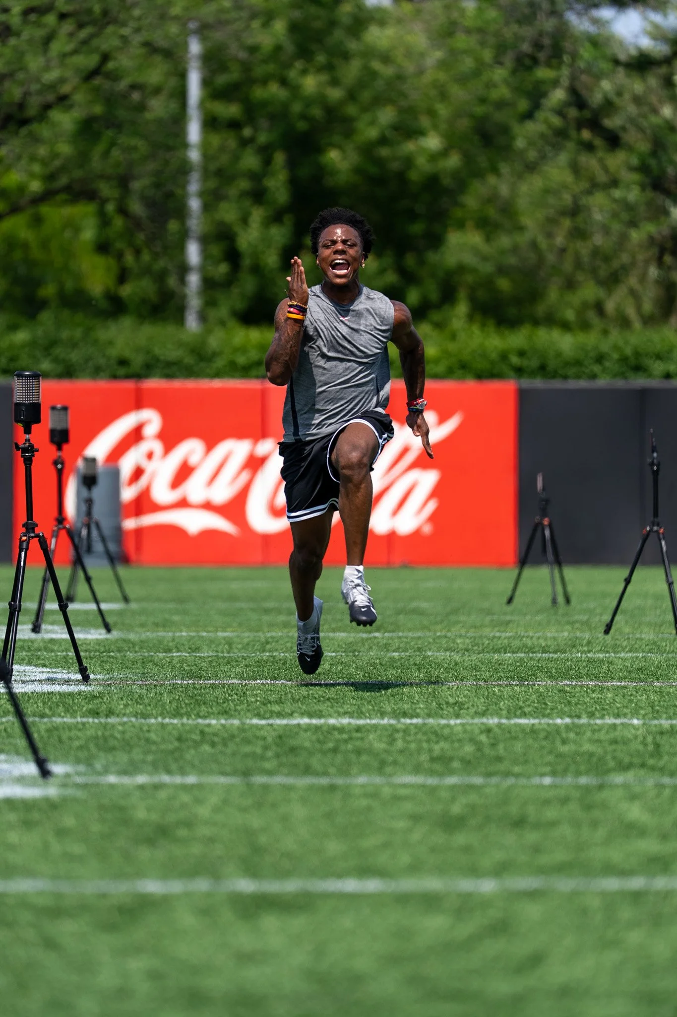 A male runner in athletic attire running on a grass field with a red advertising board behind him, set outdoors with trees in the background.