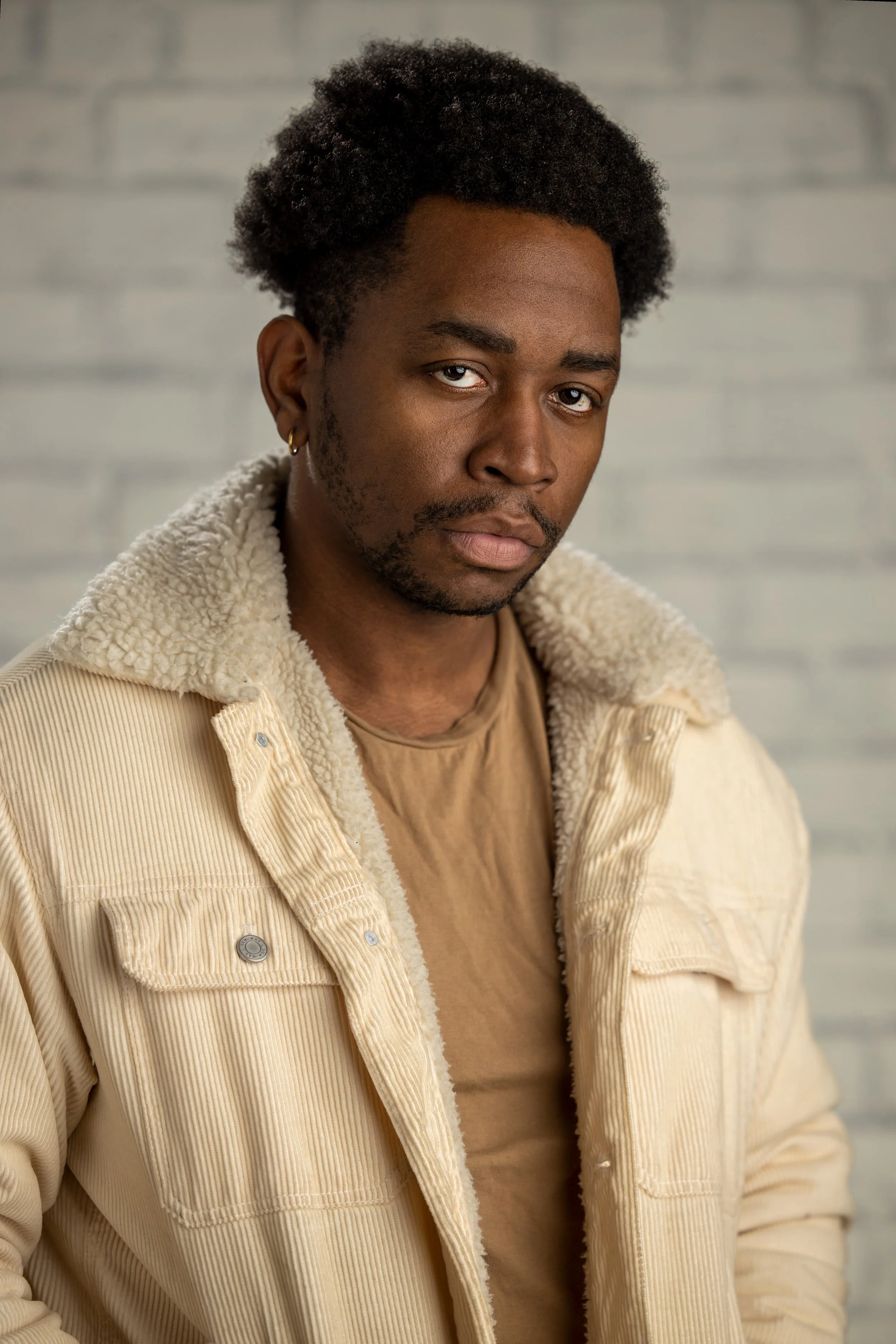 A young man with short curly hair, dark skin, and a goatee, looking at the camera with a serious expression. He is wearing a tan t-shirt and a beige corduroy jacket with a shearling collar, and has a small earring in his left ear. The background is a