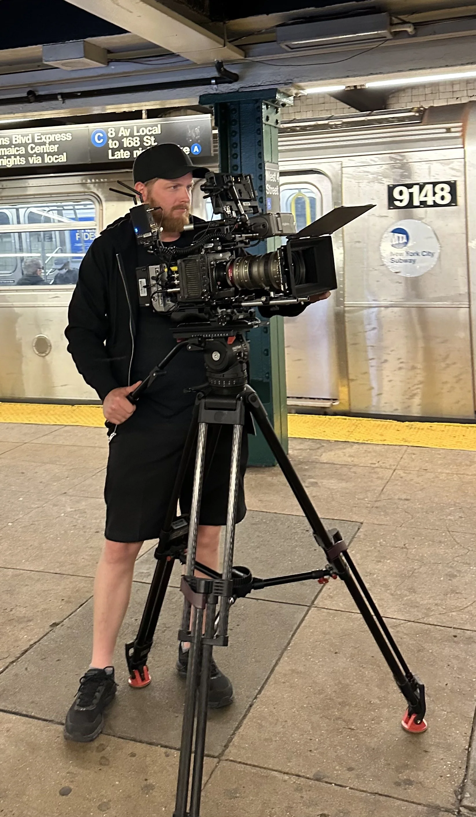 Andrew Henkelman shooting a documentary-style video at a subway station platform, with a subway train in the background and station signage overhead.