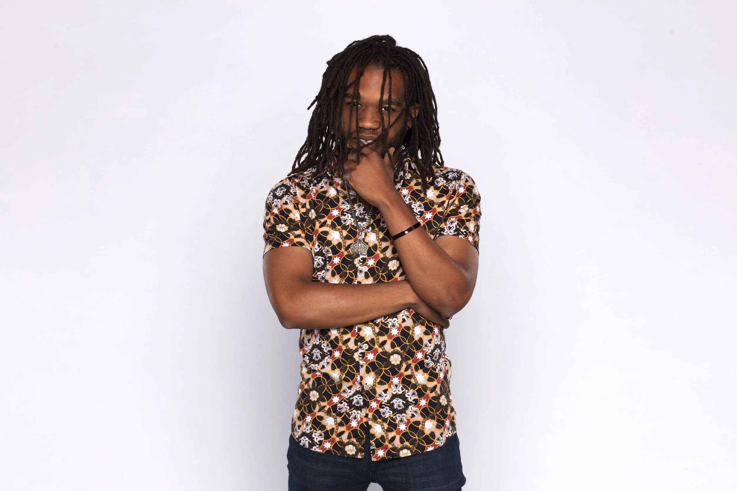 A person with long dreadlocks, wearing a colorful patterned short-sleeve shirt, standing against a plain white background.