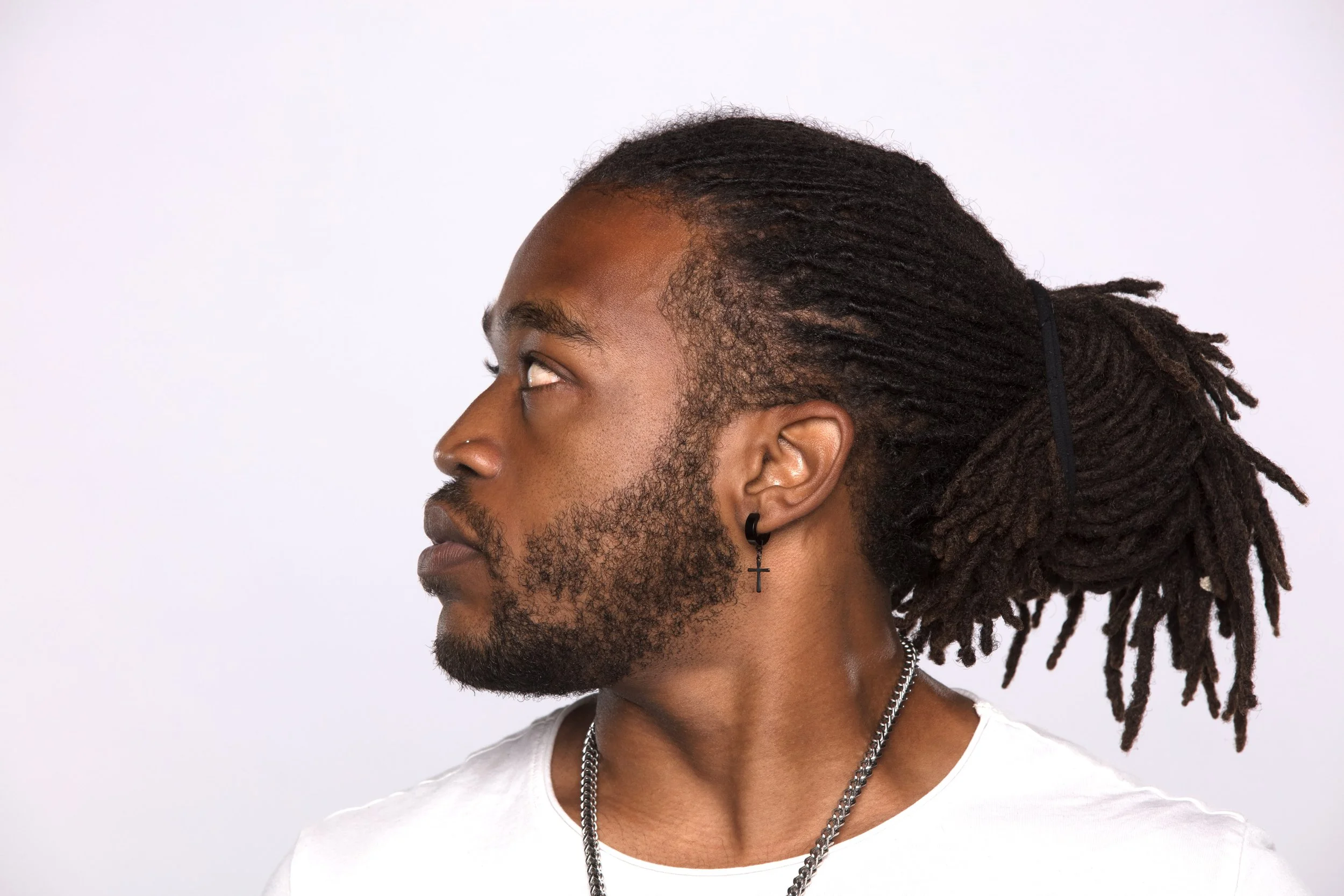 Profile of a young man with dark skin, dreadlocks, a nose piercing, wearing a cross earring, a silver chain, and a white shirt against a white background.