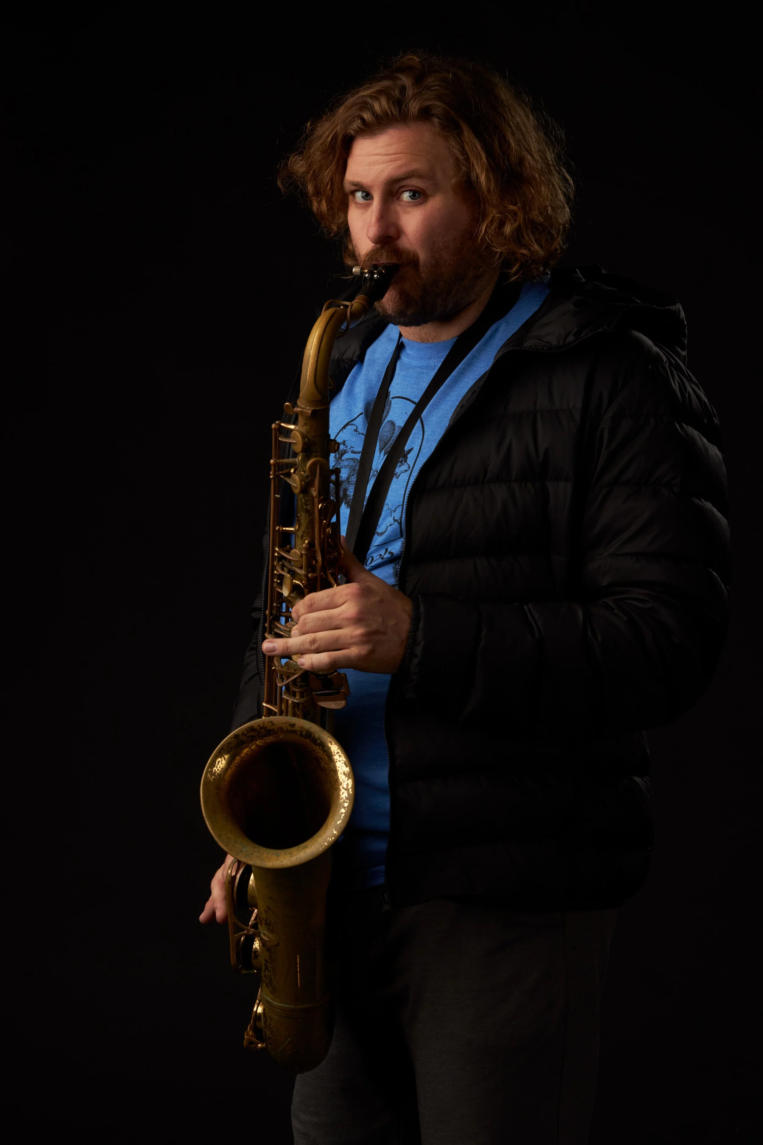 A man with curly reddish-brown hair and facial hair playing a saxophone against a black background. He is wearing a black jacket over a blue t-shirt.