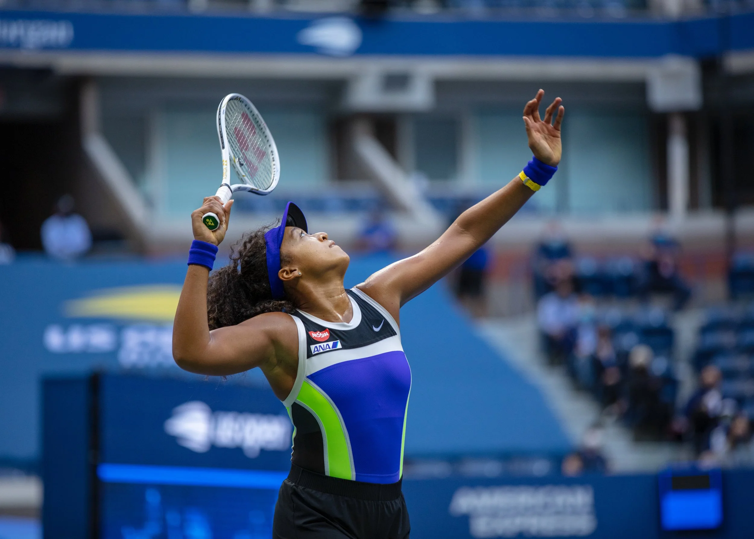 A photograph by Andrew Henkelman of Naomi Osaka preparing to serve during a match, wearing her iconic 2018 blue visor, a sports dress with blue, yellow, and black accents, and wristbands, on a the main court of the  with spectators in the background.