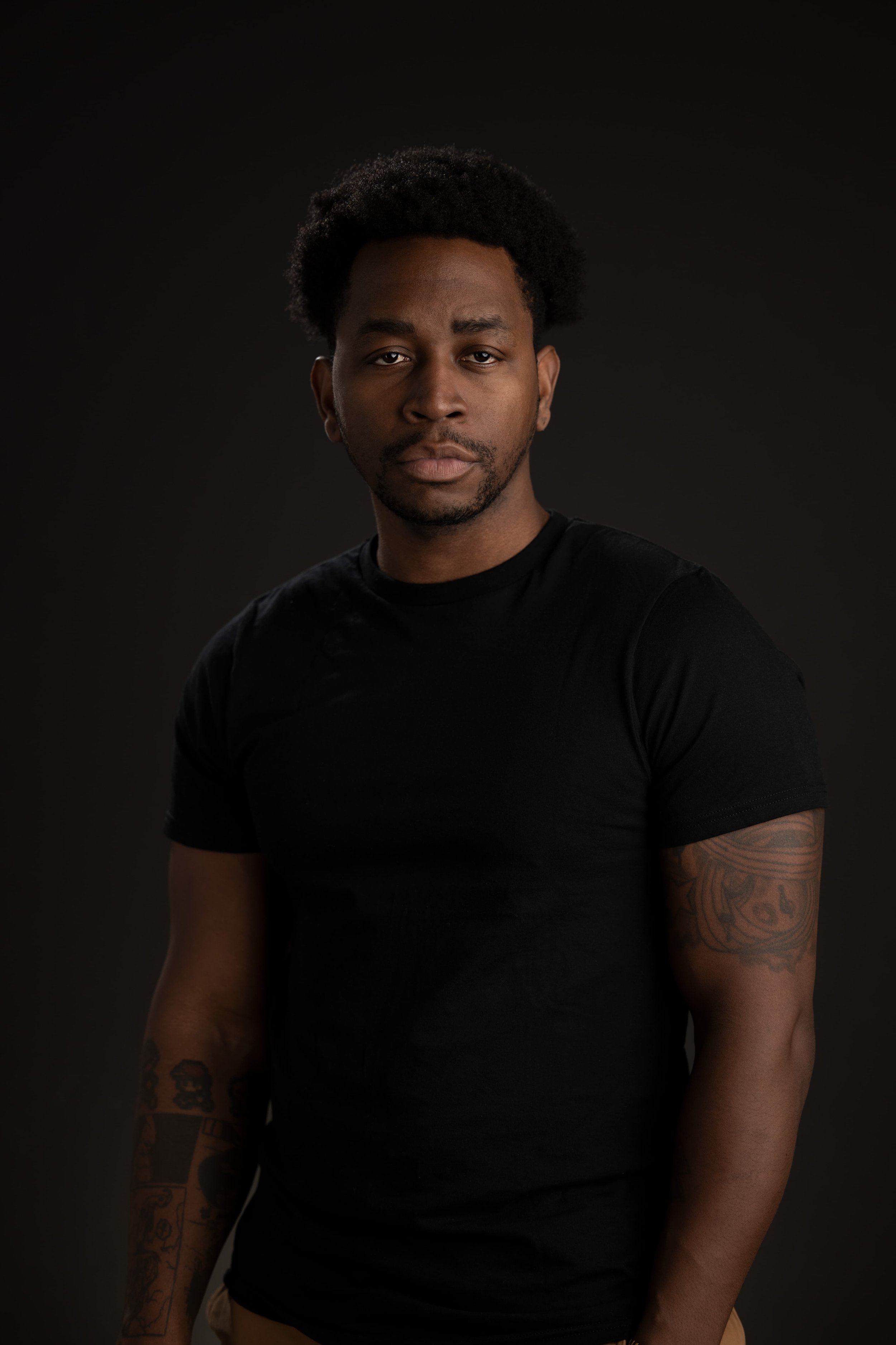 Portrait of a young man with dark hair, facial hair, tattoos on his arms, wearing a black t-shirt, against a dark background.