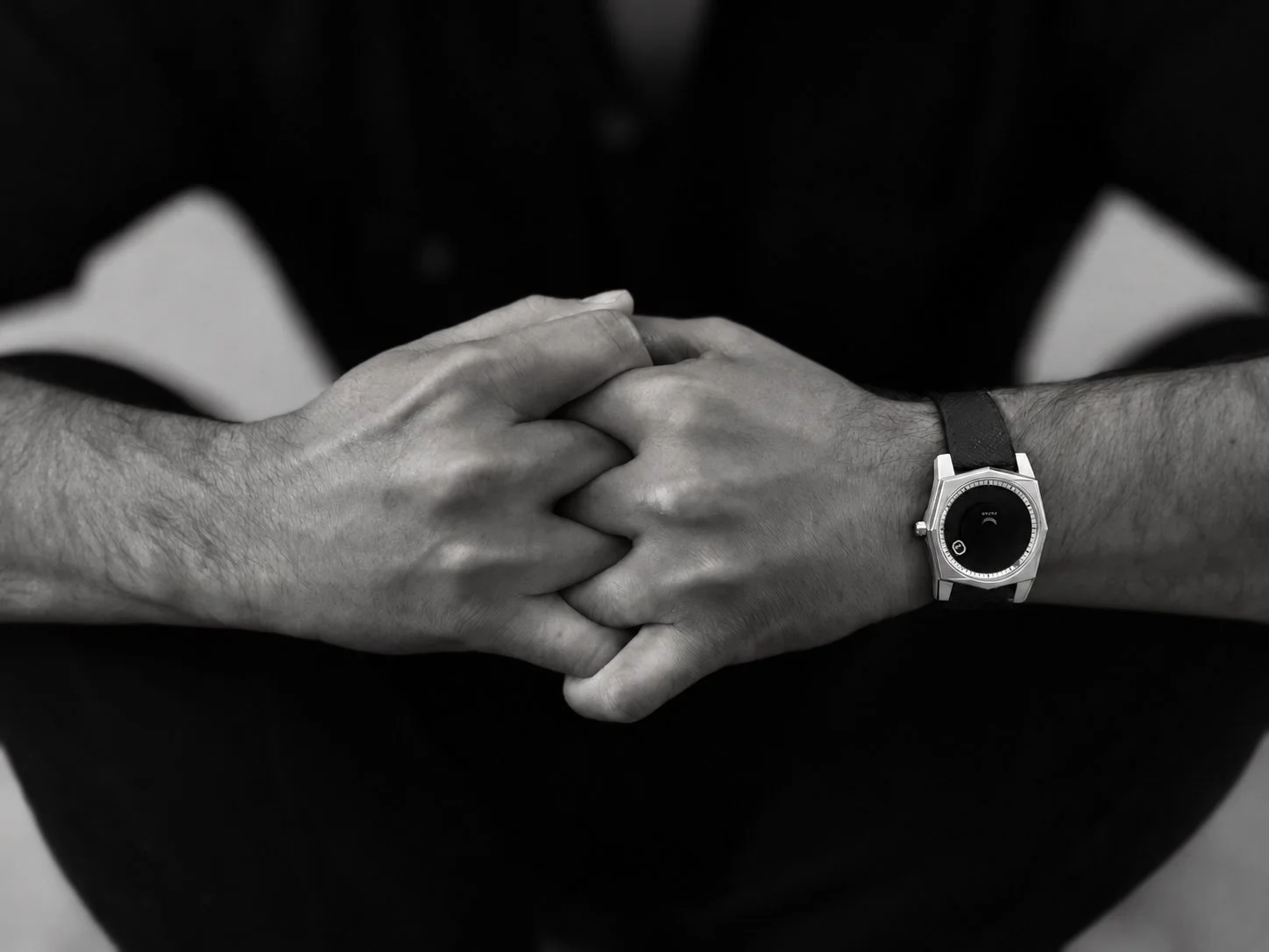 Black and white photo of two hands clasped together, with one wearing a modern wristwatch, against a dark background.