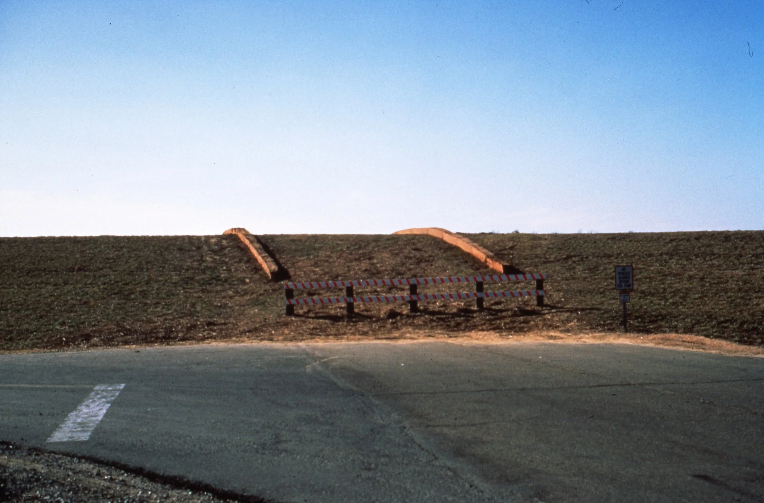 A road with a barricade blocking access to a grassy hillside. There are two brick steps leading up the hill, and a sign on the right side.