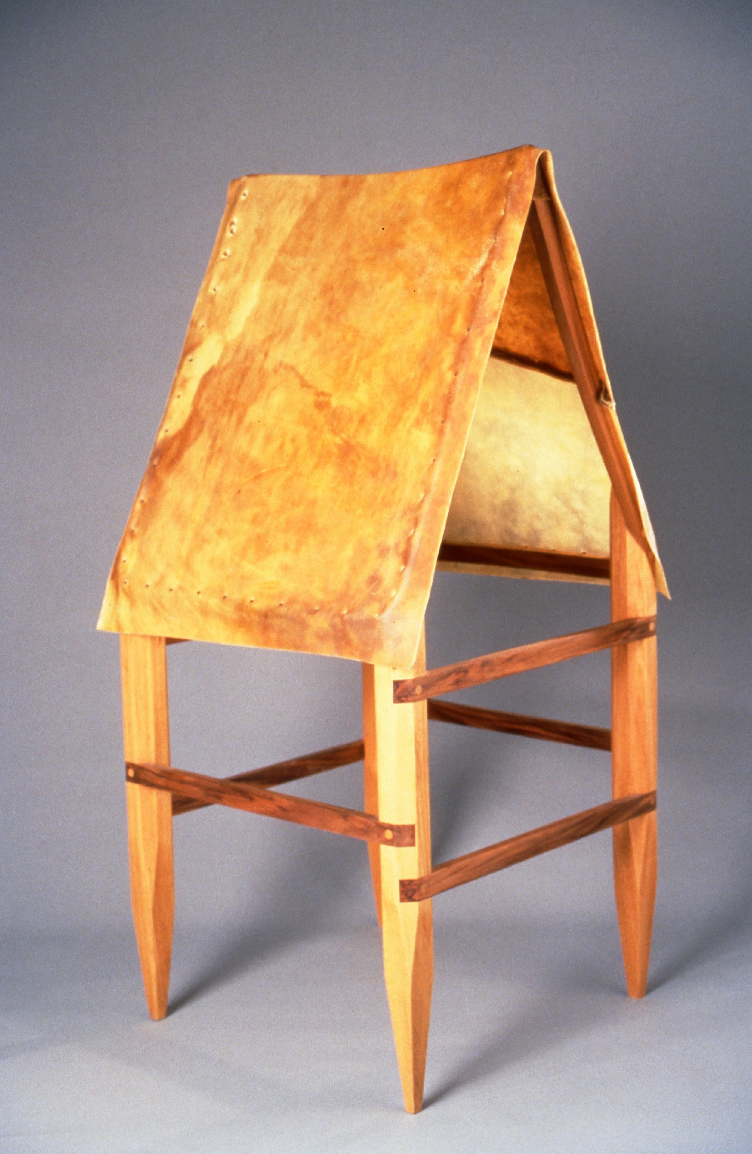 A wooden art piece resembling a small A-frame house with a textured, rust-colored roof and a small interior shelf, set against a plain gray background.