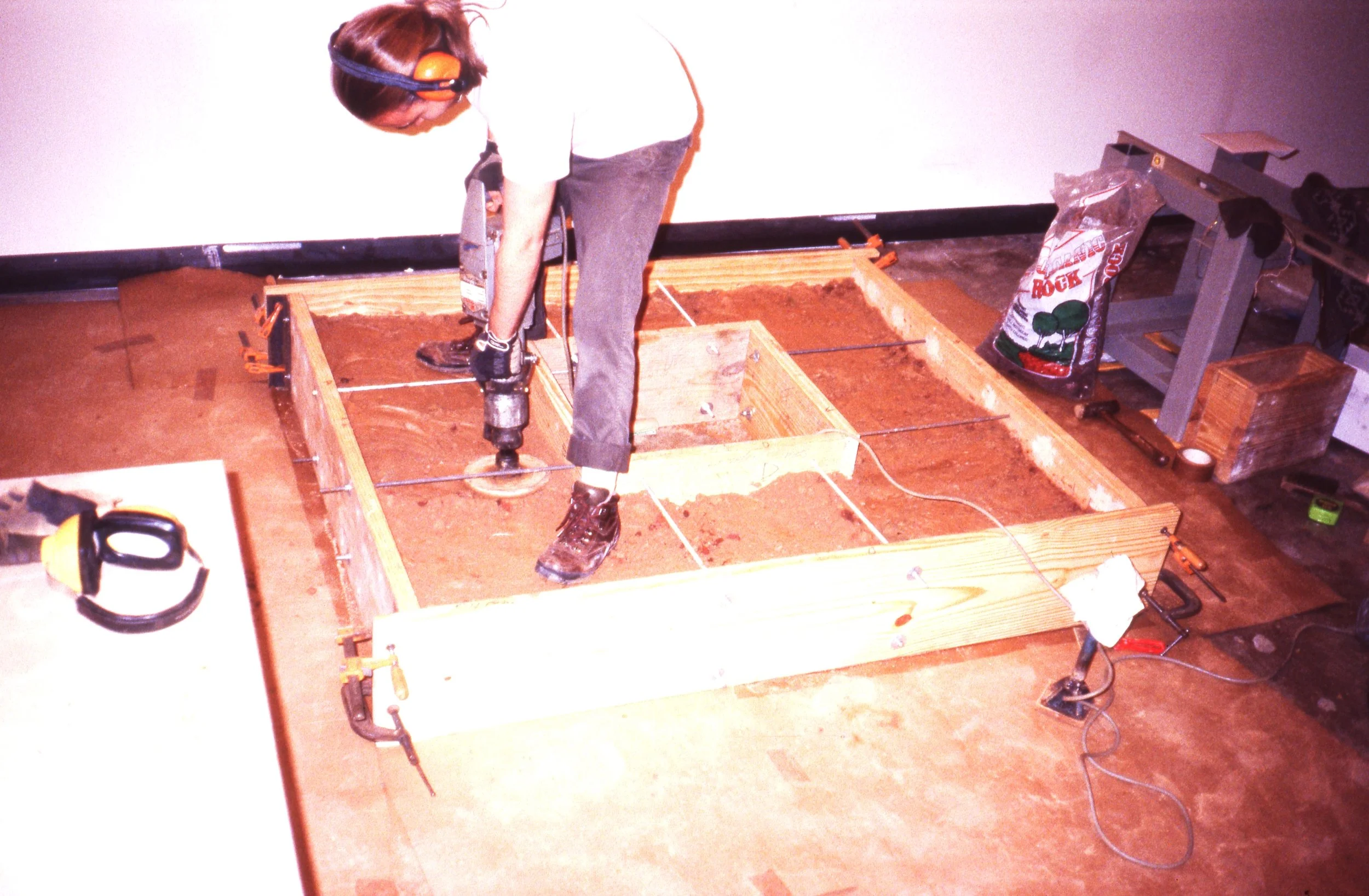 Person using a power drill to work on a wooden frame construction on the floor, with tools and construction materials around.