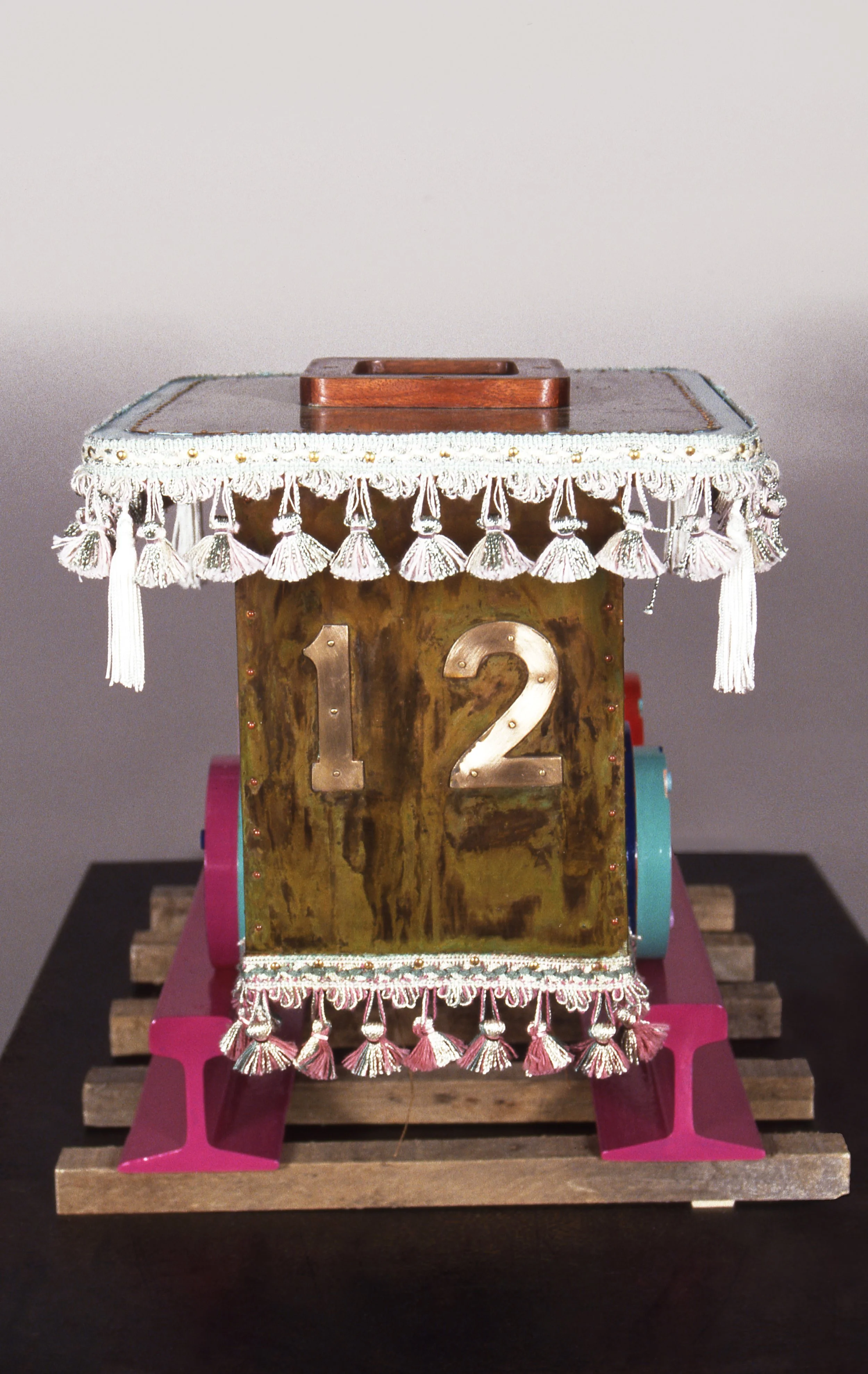 Decorative wooden block with the number 12, topped with a fringed fabric cover, standing on pink and wooden bases.