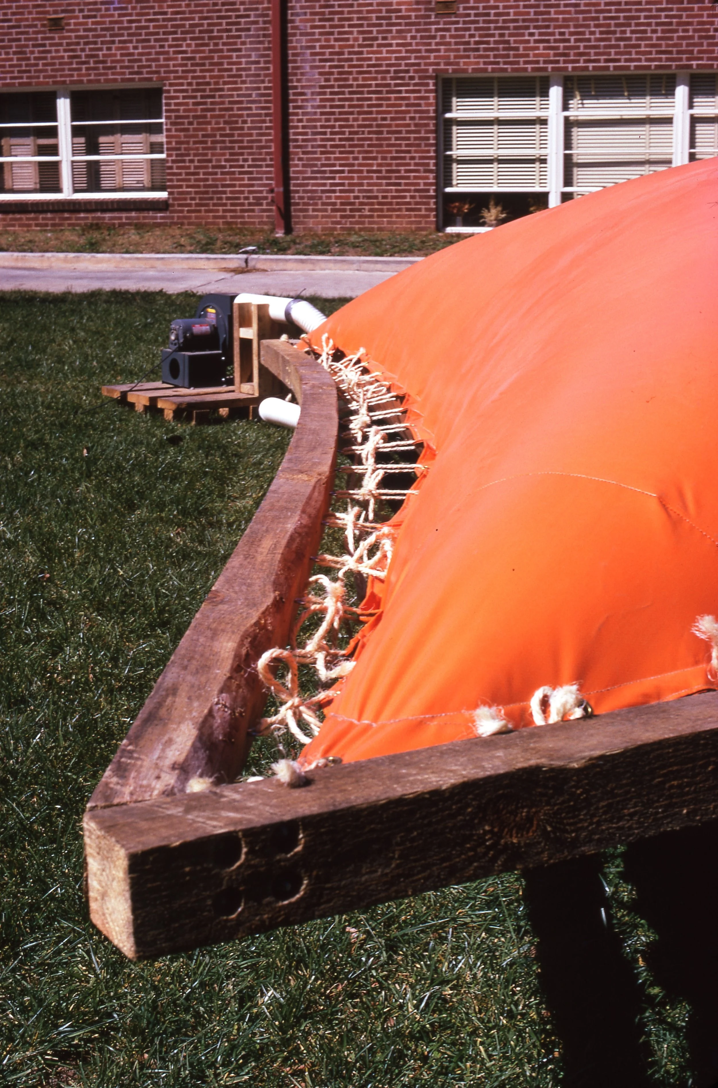 Close-up of an orange weather balloon tethered to a wooden frame, with a black device and white pipes in the background, on a grassy area near a brick building.