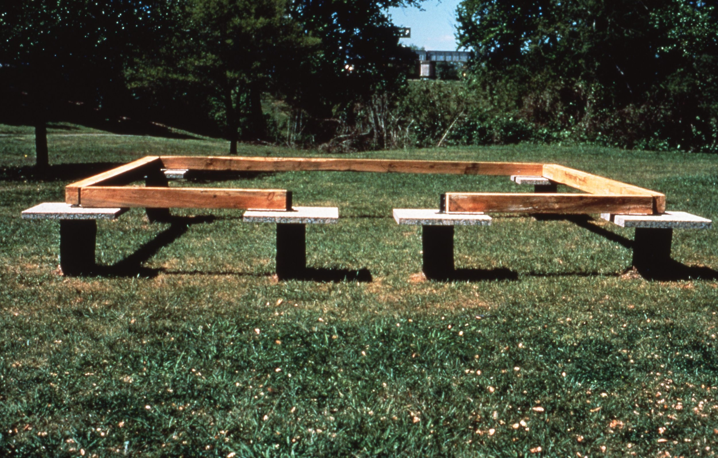 A park bench frame under construction in a grassy park with trees and a structure in the background.