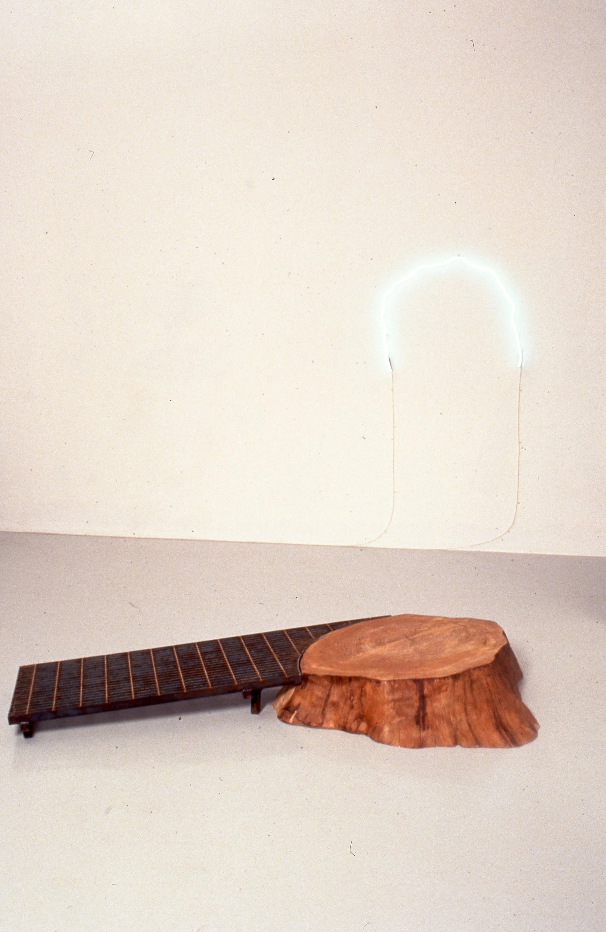A sculptural art piece resembling a guitar, made from a piece of wood for the body and a dark wooden or metal section for the neck, placed on a light-colored surface against a white wall.