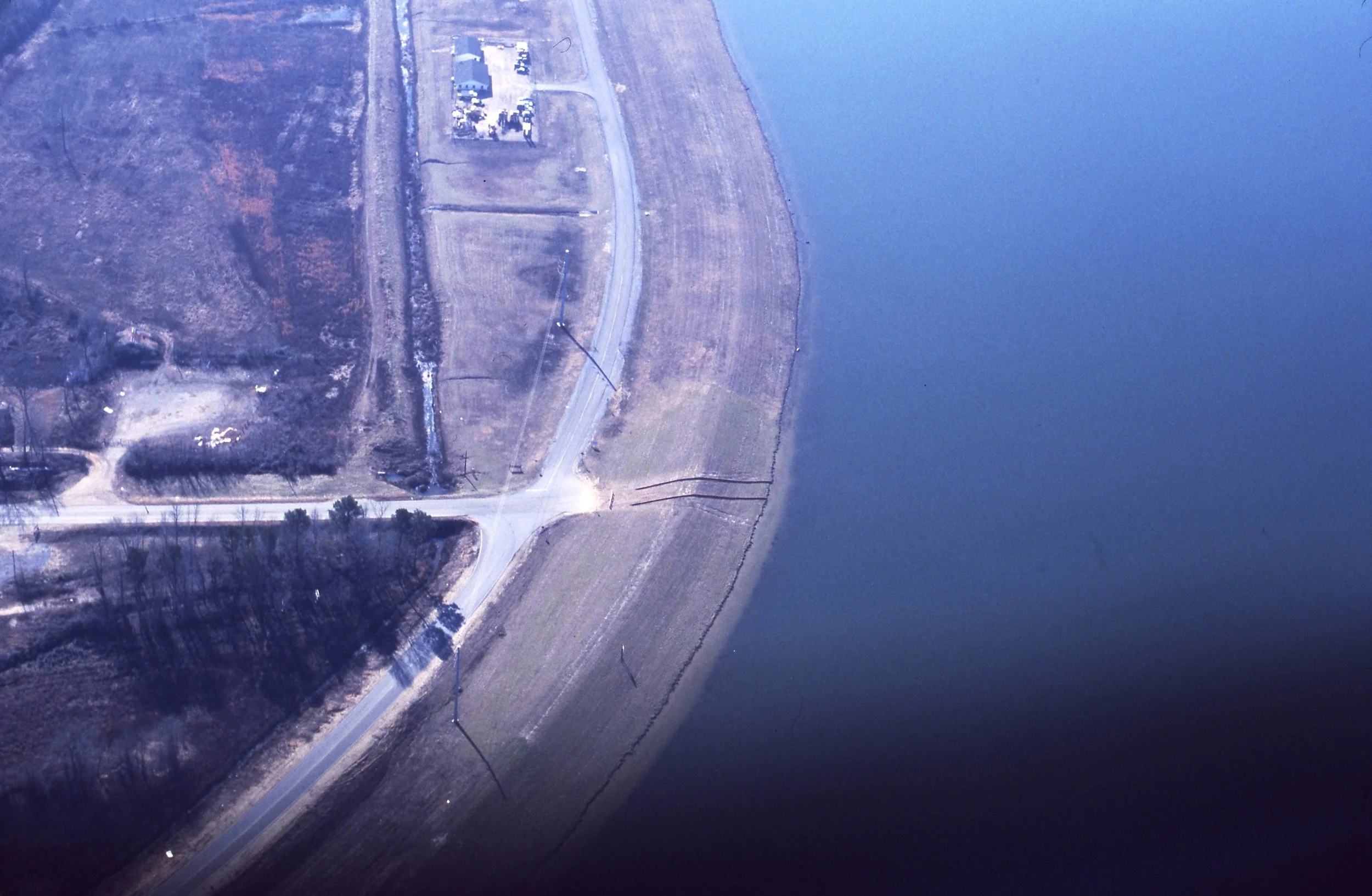 Aerial view of a rural landscape with a body of water on the right, a road intersecting a curved road, and an open field with some buildings and trees.