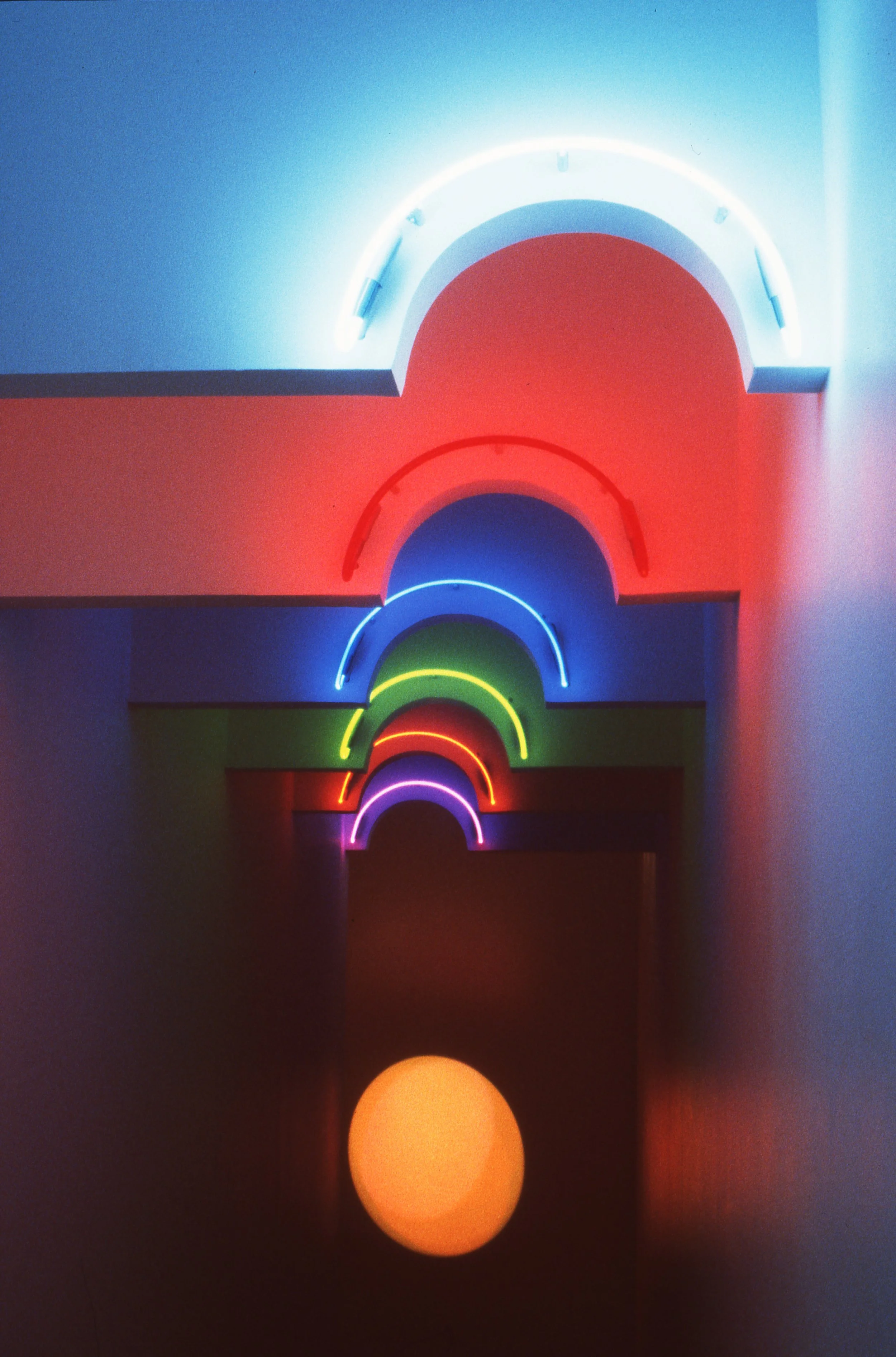 Multiple illuminated neon arches in rainbow colors, stacked vertically against a wall, with a small glowing orange sphere on the floor reflecting the lights.