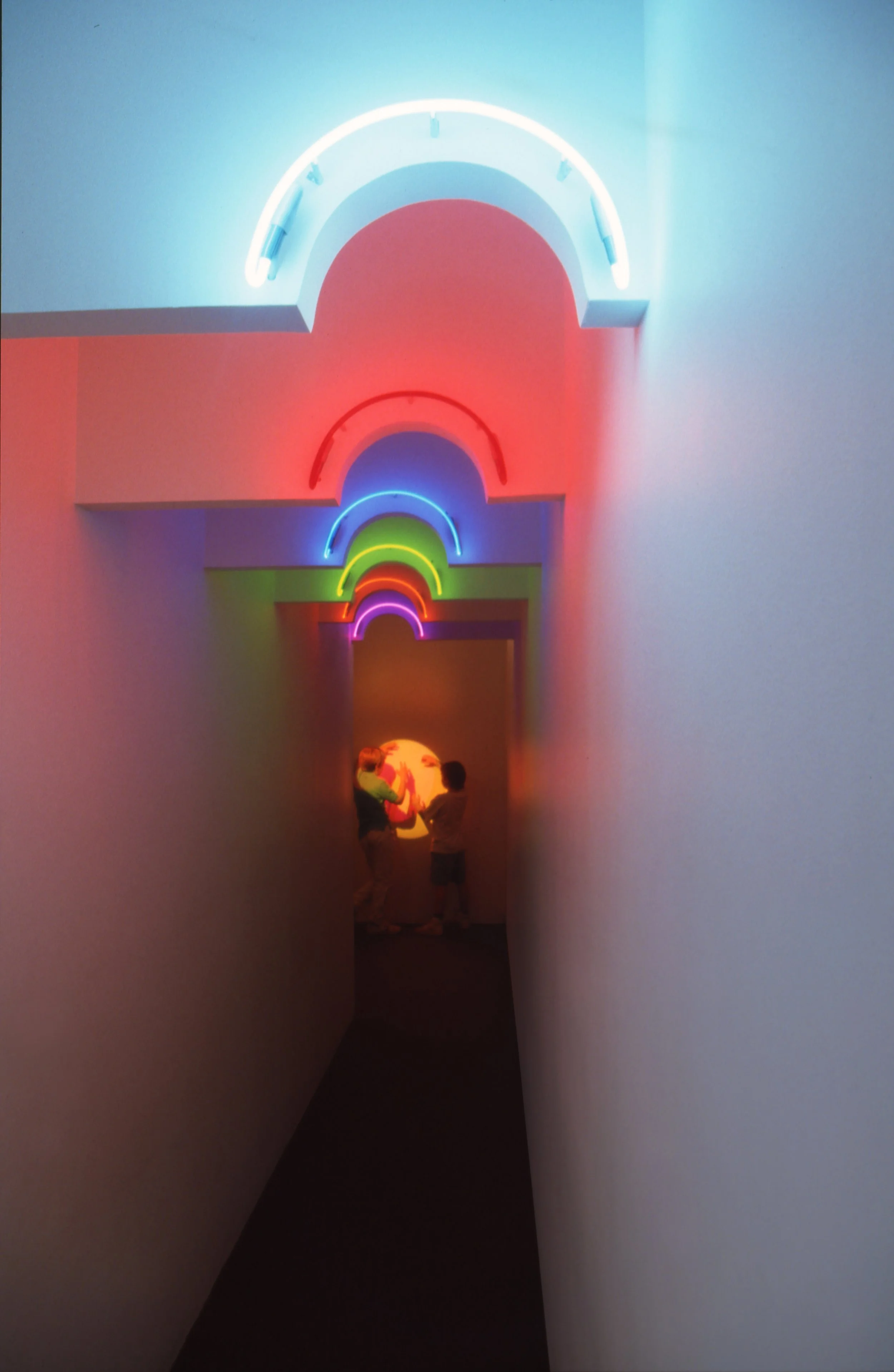 A hallway with colorful neon rainbow lights on the ceiling, leading to an art installation where two children and an adult interact with a glowing, spherical object.