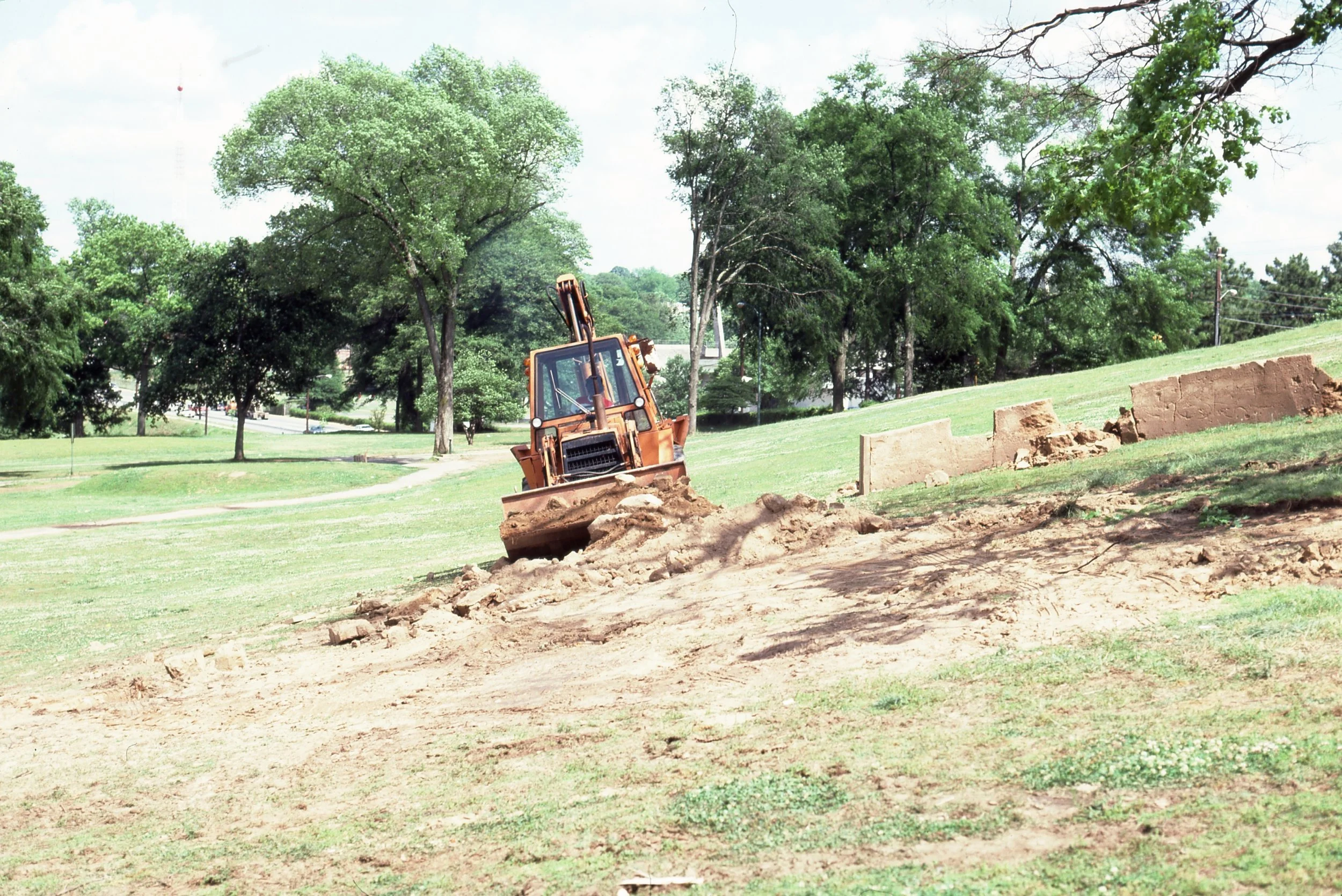 A bulldozer clearing away dirt on a grassy park with trees in the background.