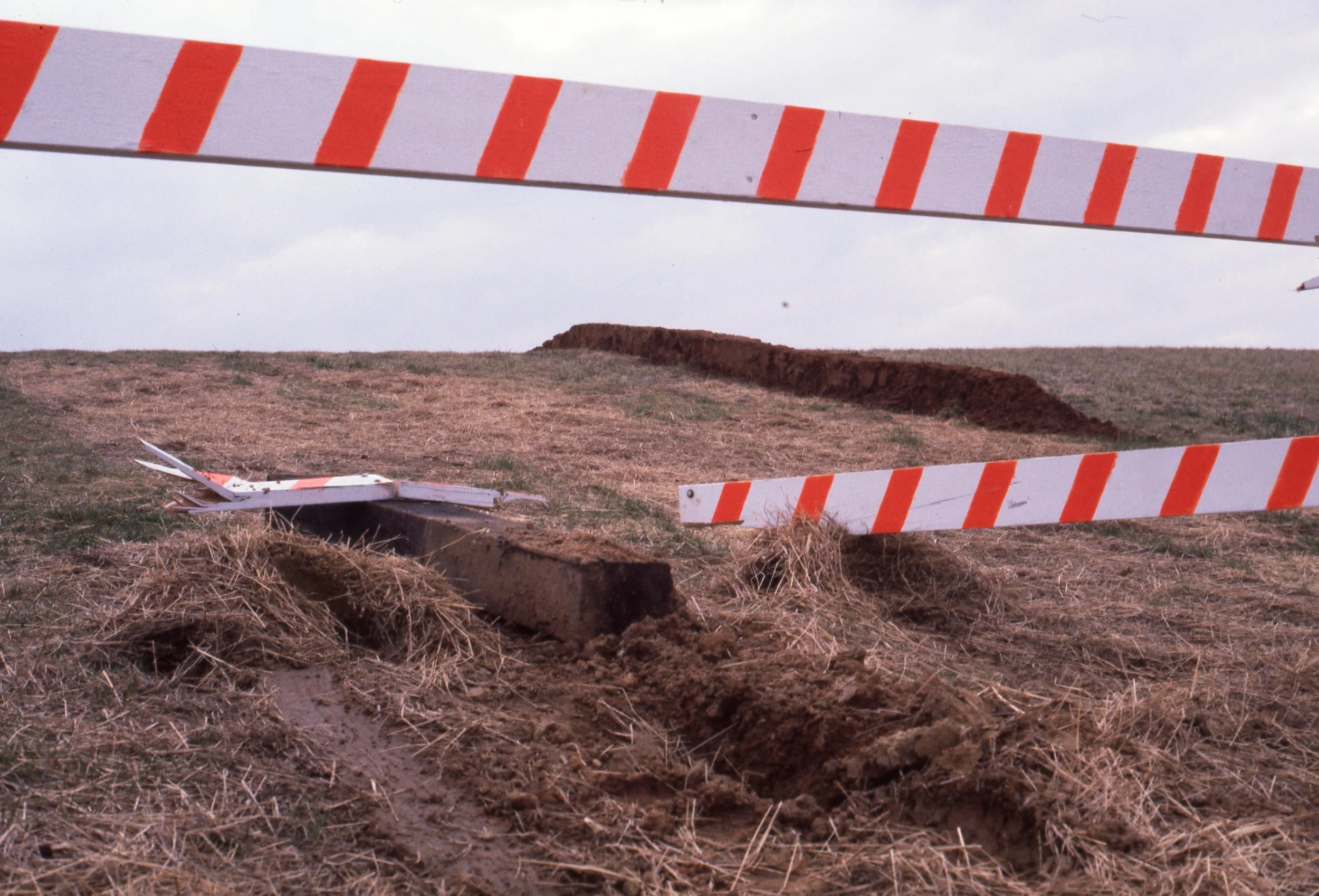 A damaged construction barrier fallen over a dirt road with exposed dirt and grass in an open field.