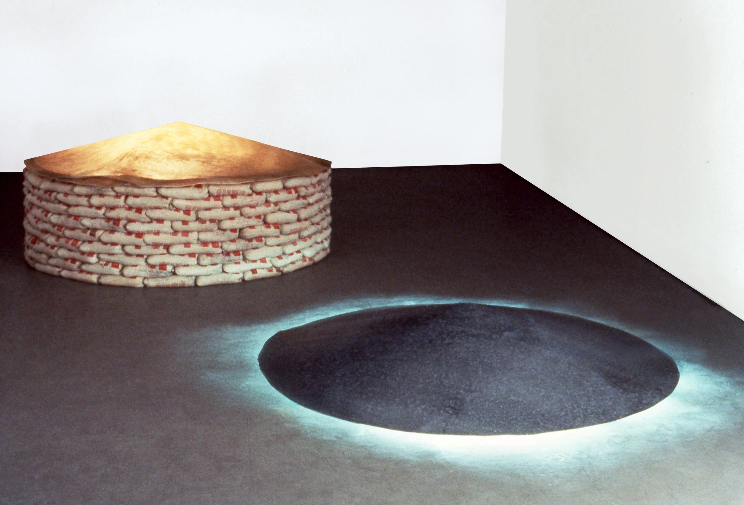 Contemporary art installation featuring a woven, circular object resembling a stack of fabric or paper, placed on a gray floor against a white wall, with an illuminated black oval shape on the floor.