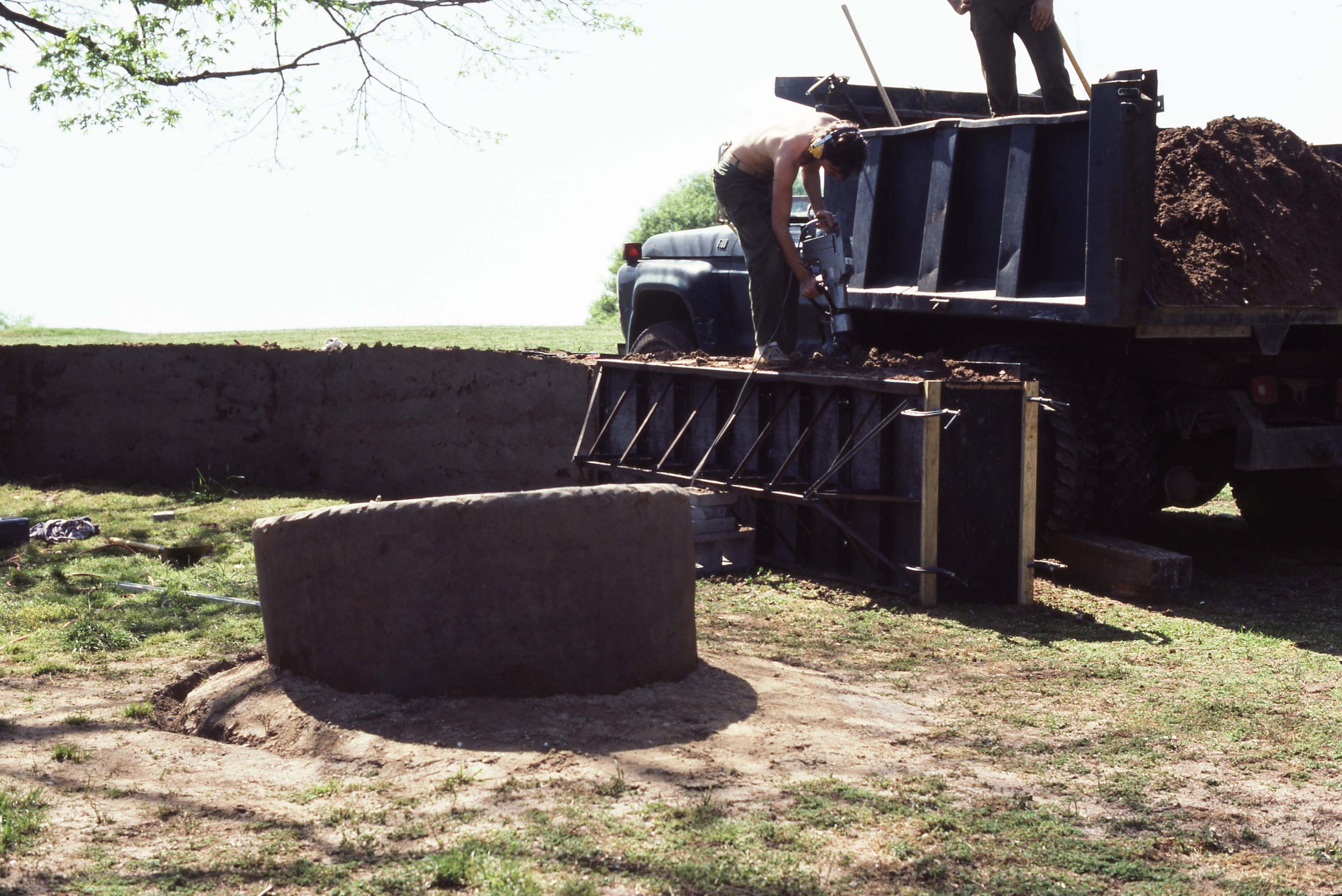A man is working on a construction site, using a power tool on a large concrete well curb. There is a truck loaded with dirt nearby and a person standing on the truck bed. The area is outdoors with grass and a tree branch visible overhead.