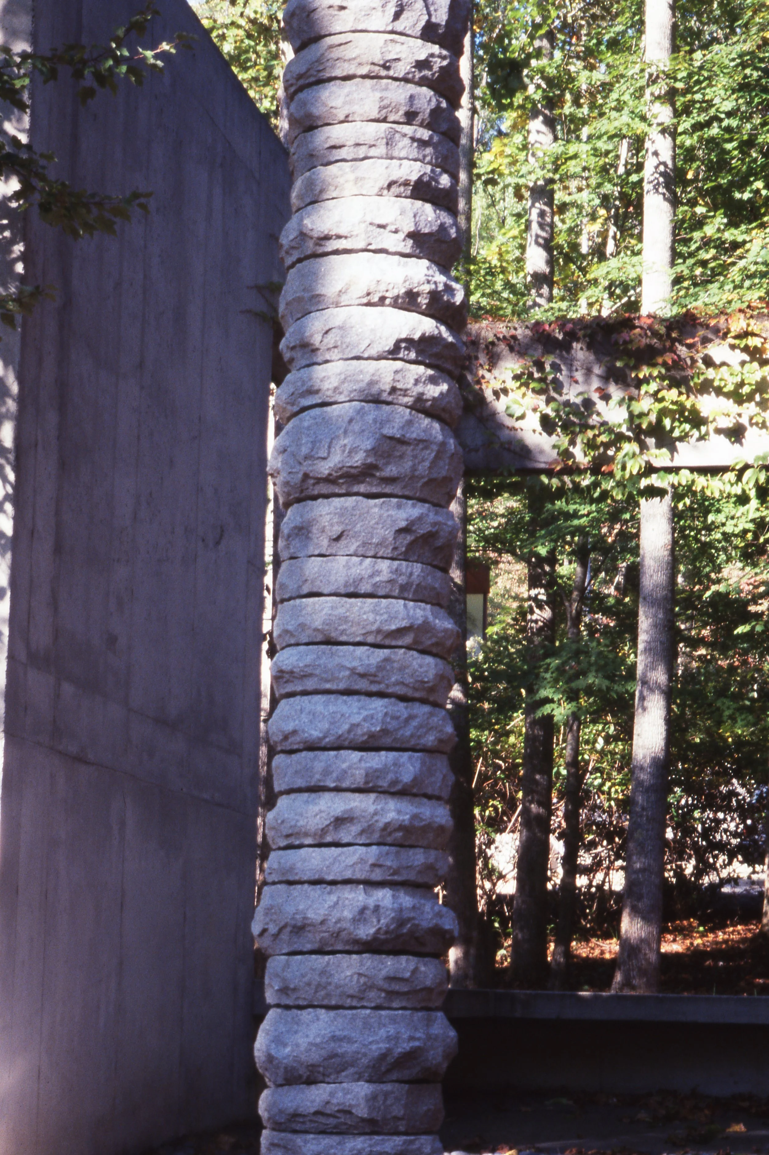 A stone pillar with stacked, irregularly shaped rocks standing next to a concrete wall, with trees and foliage in the background.