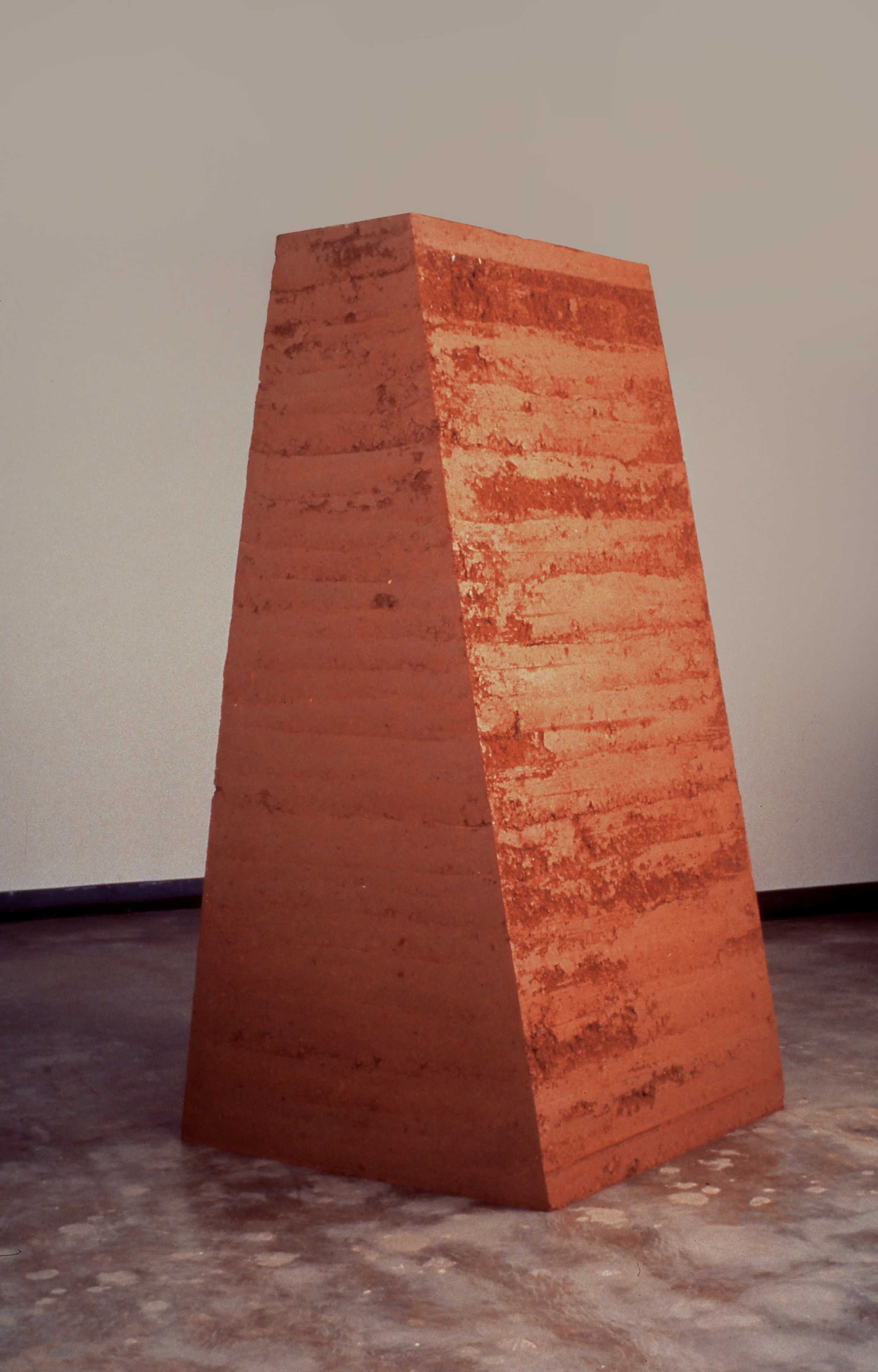 A tall, trapezoidal sculpture made of reddish-brown textured material, situated indoors on a concrete floor with a plain light-colored wall in the background.