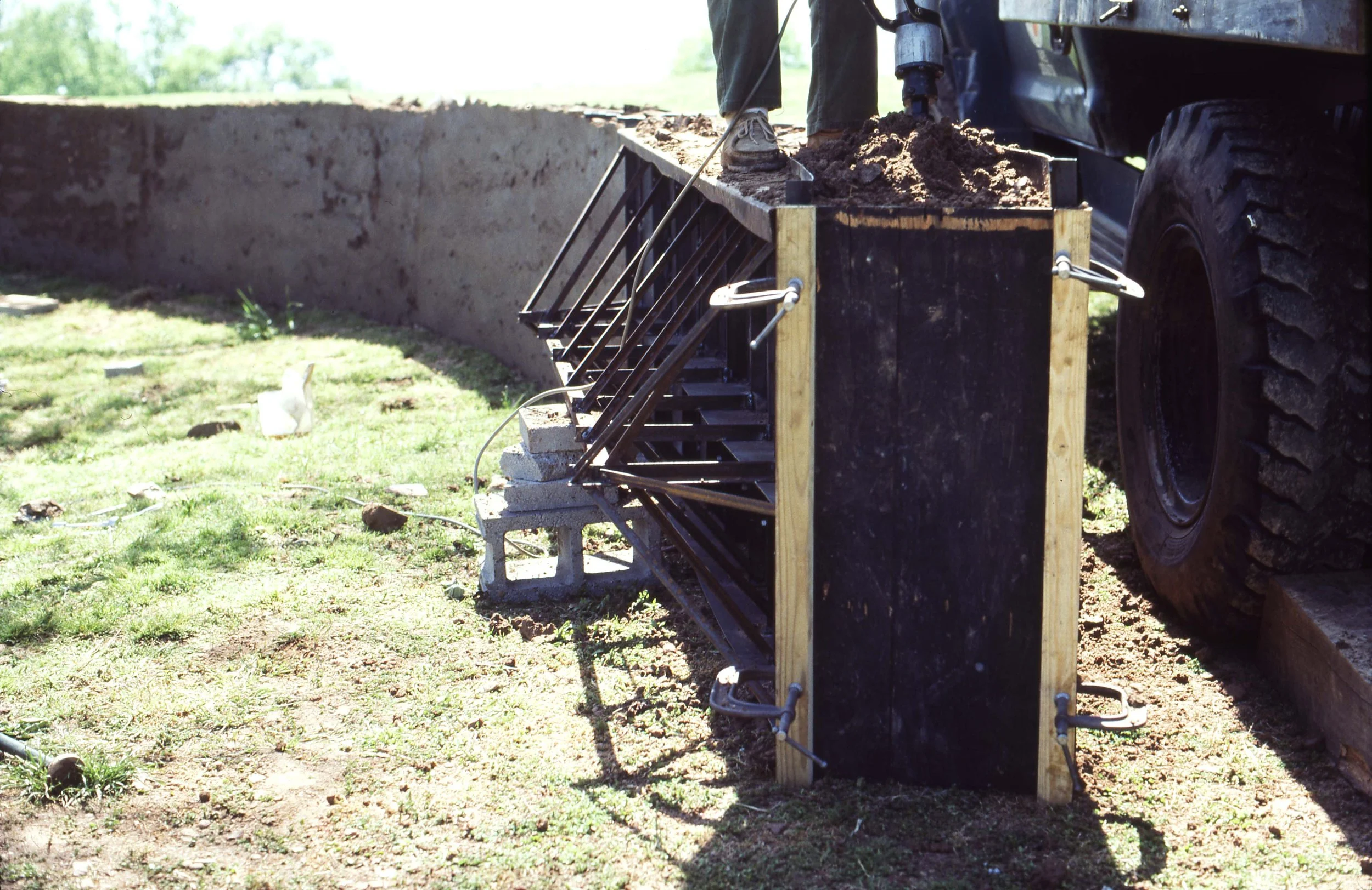 A construction site with a close-up of a black and Wooden formwork frame for concrete pouring. A worker's feet are visible standing on the formwork. The background shows a grassy area and a clear sky.