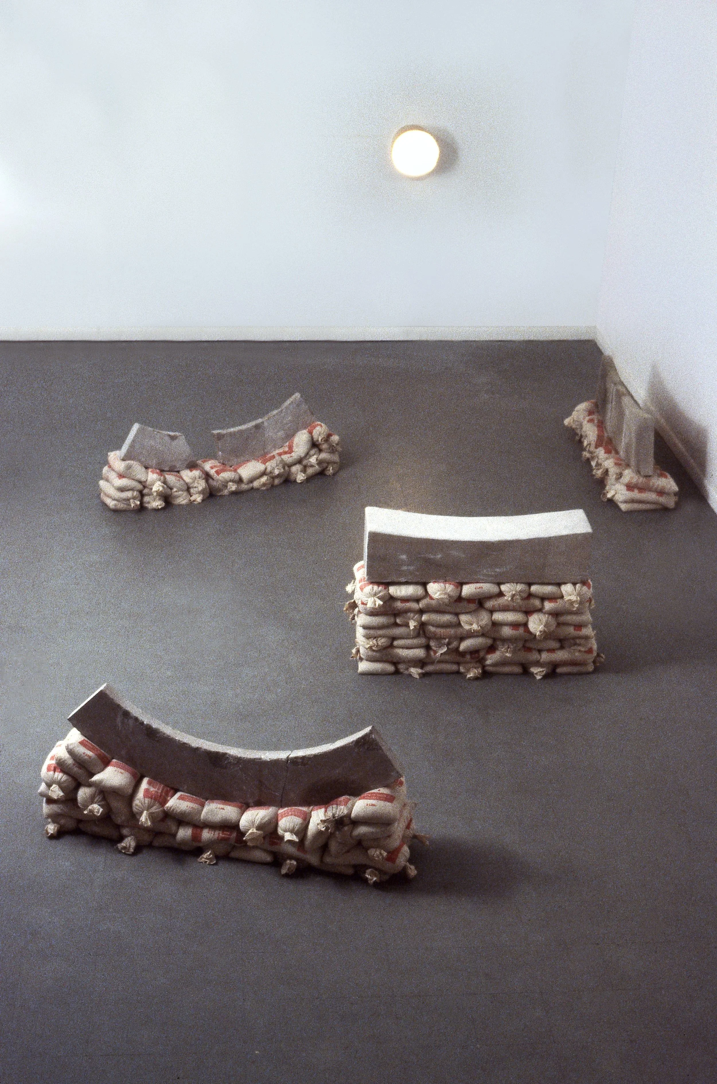 Abstract art installation with curved and rectangular forms made of layered bags or fabrics, arranged on a dark floor in a room with white walls and a ceiling light.