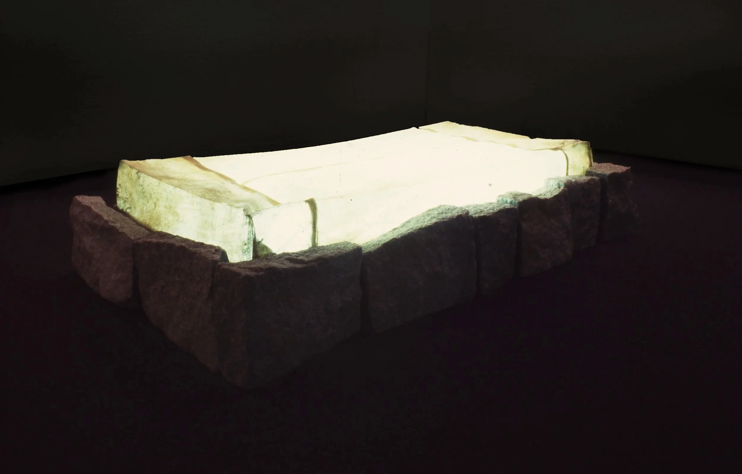 Illuminated ancient rectangular stone basin or vessel, surrounded by rough stone blocks, set against a dark background.