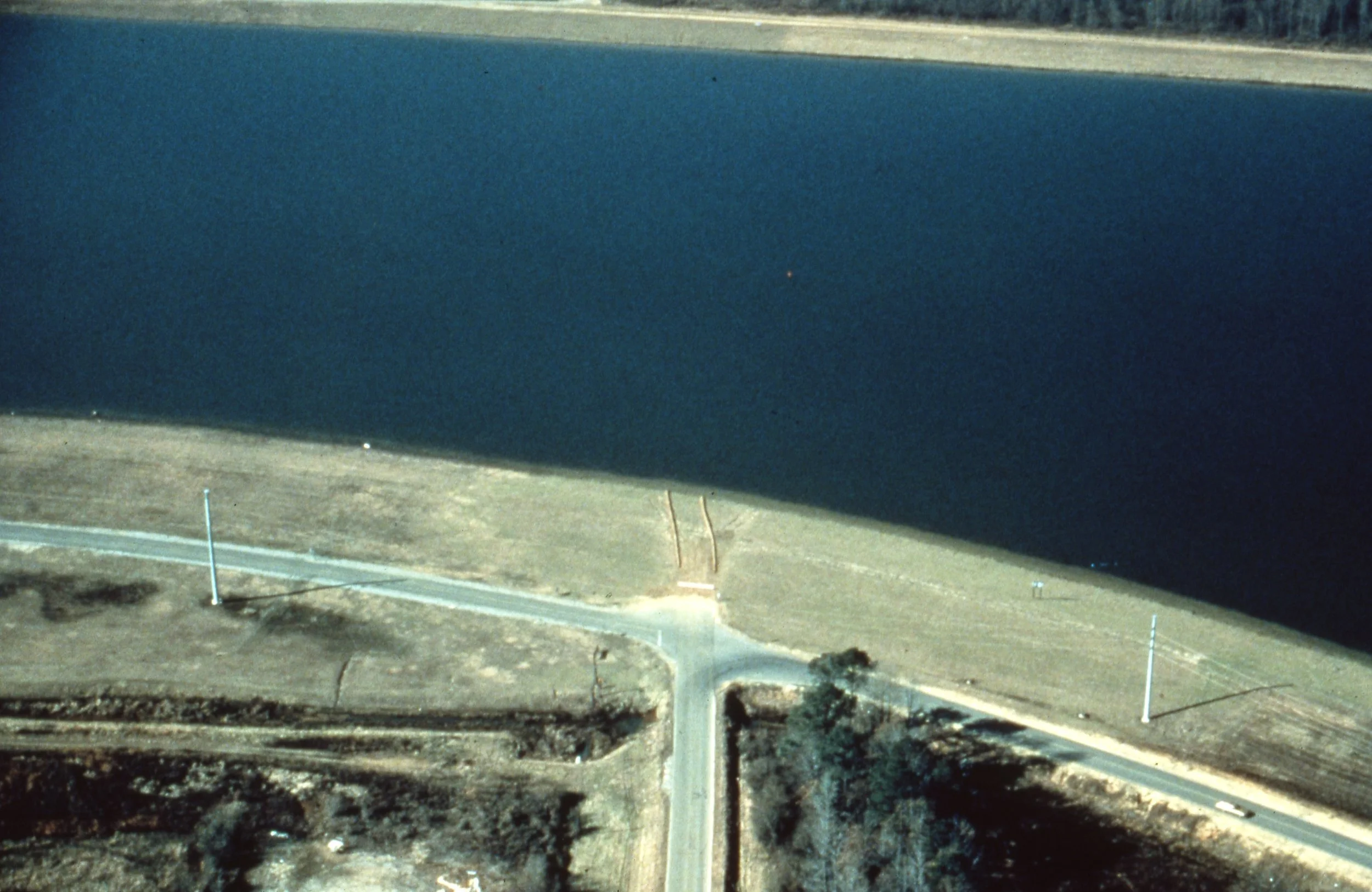 Aerial view of a rural area with a large body of water, a small road crossing the land, and some trees and open fields.
