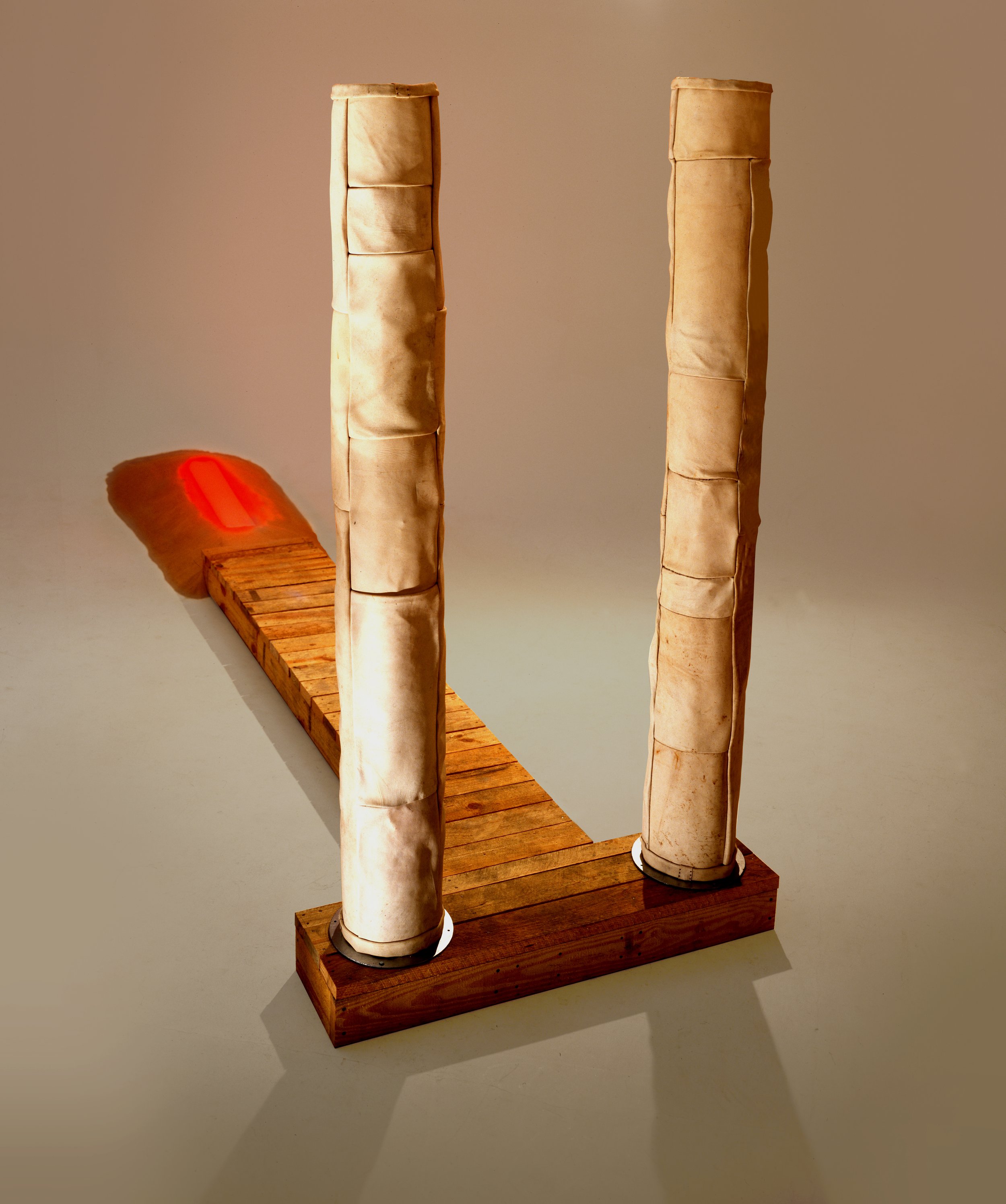 Art installation featuring two tall, vertical paper-like structures standing on a wooden base with a wooden walkway and a red light projection casting a shadow.
