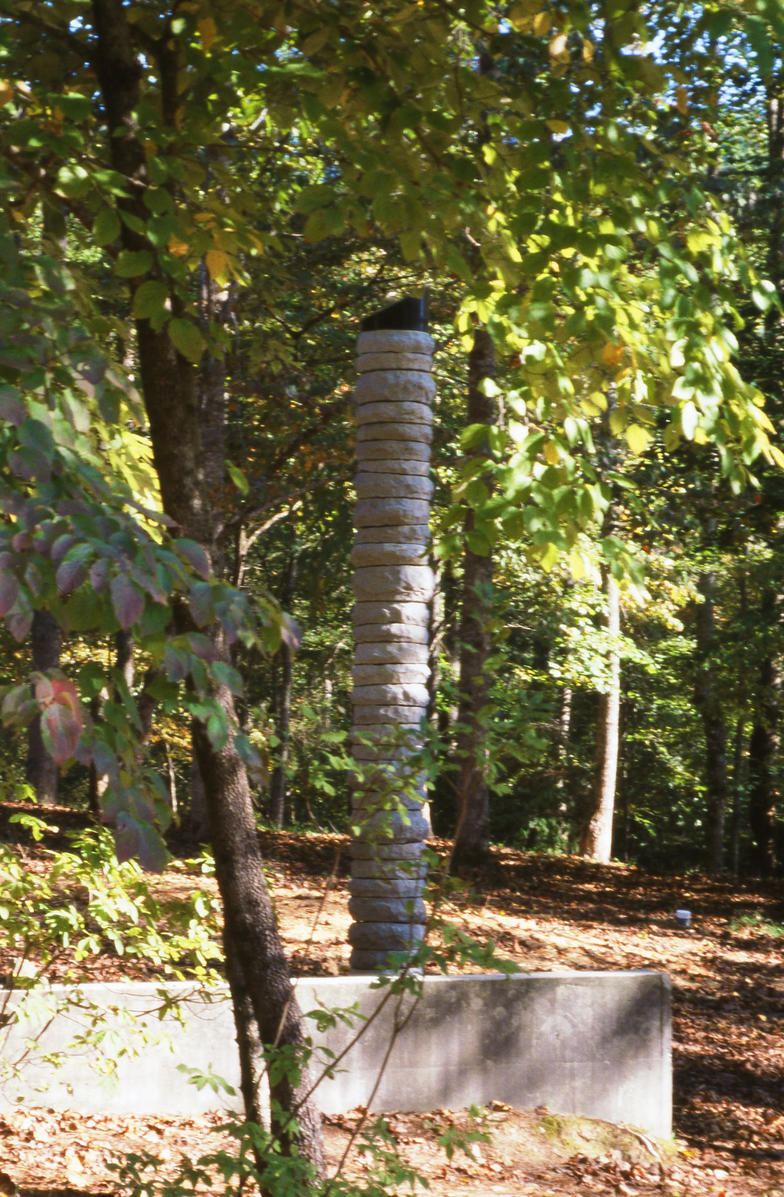 A tall, narrow stone sculpture resembling a totem pole stands among trees in a forested area, with sunlight filtering through the leaves.