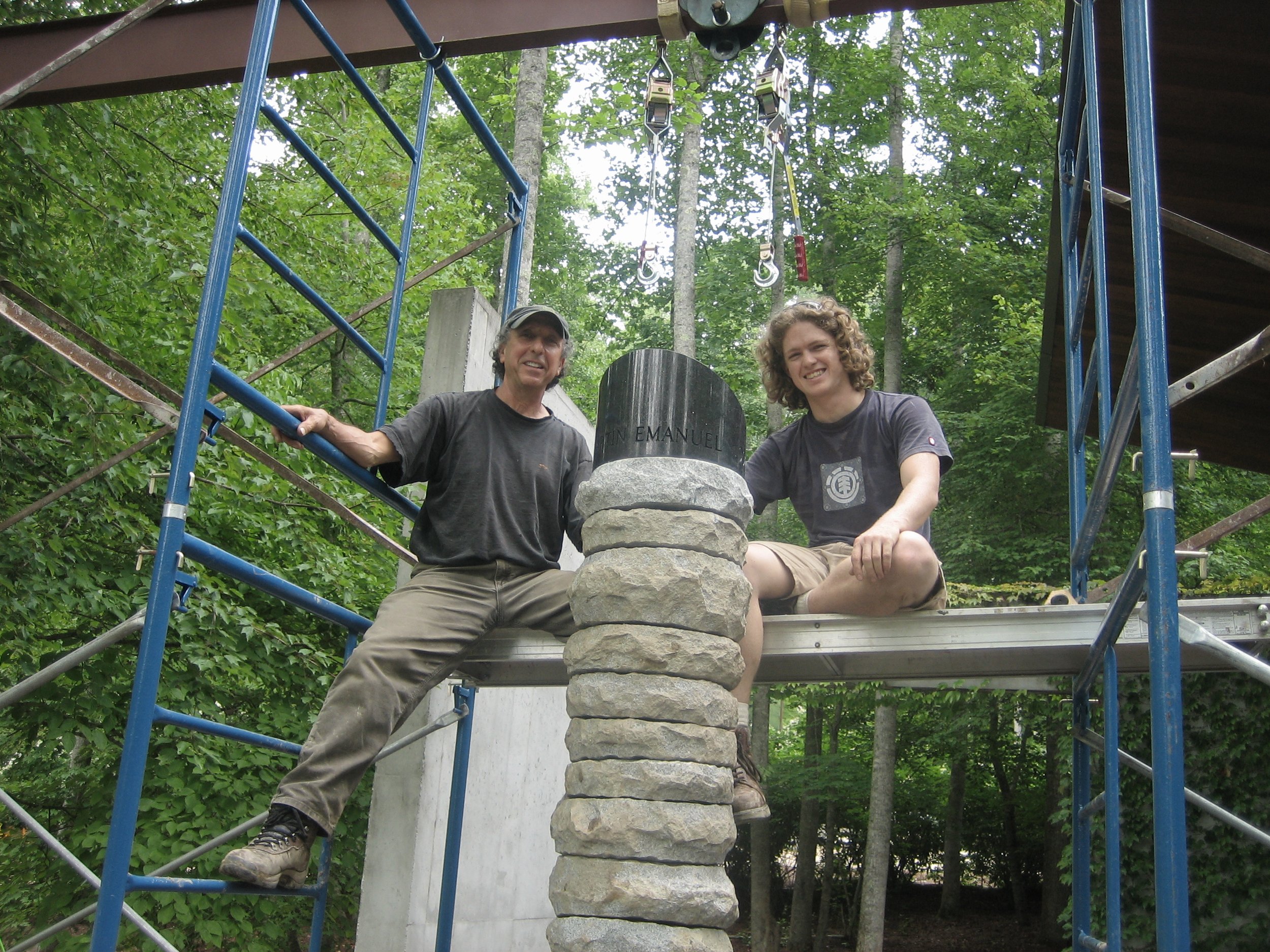 Two men sitting on a metal structure in a wooded area with a stone monument between them. One man is wearing a black shirt and a cap, and the other is wearing a gray shirt with curly hair. They are smiling and appear to be working on or celebrating a