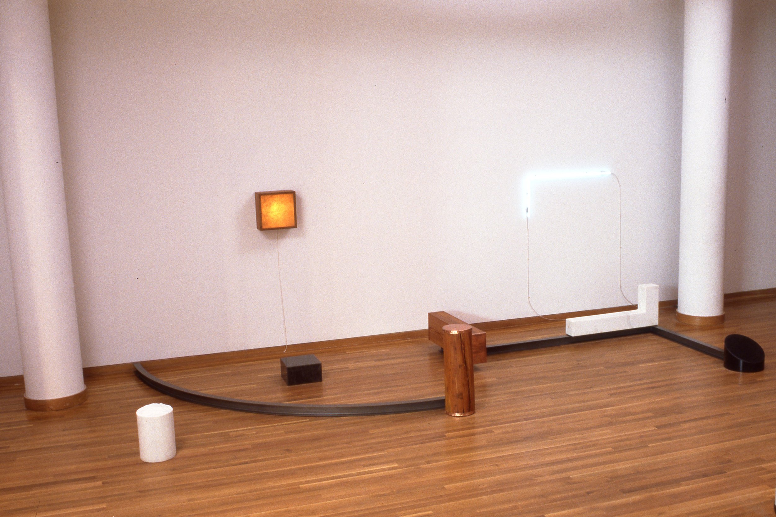 Abstract mixed-media art installation with wooden, metal, and foam elements on a wooden floor against a white wall.